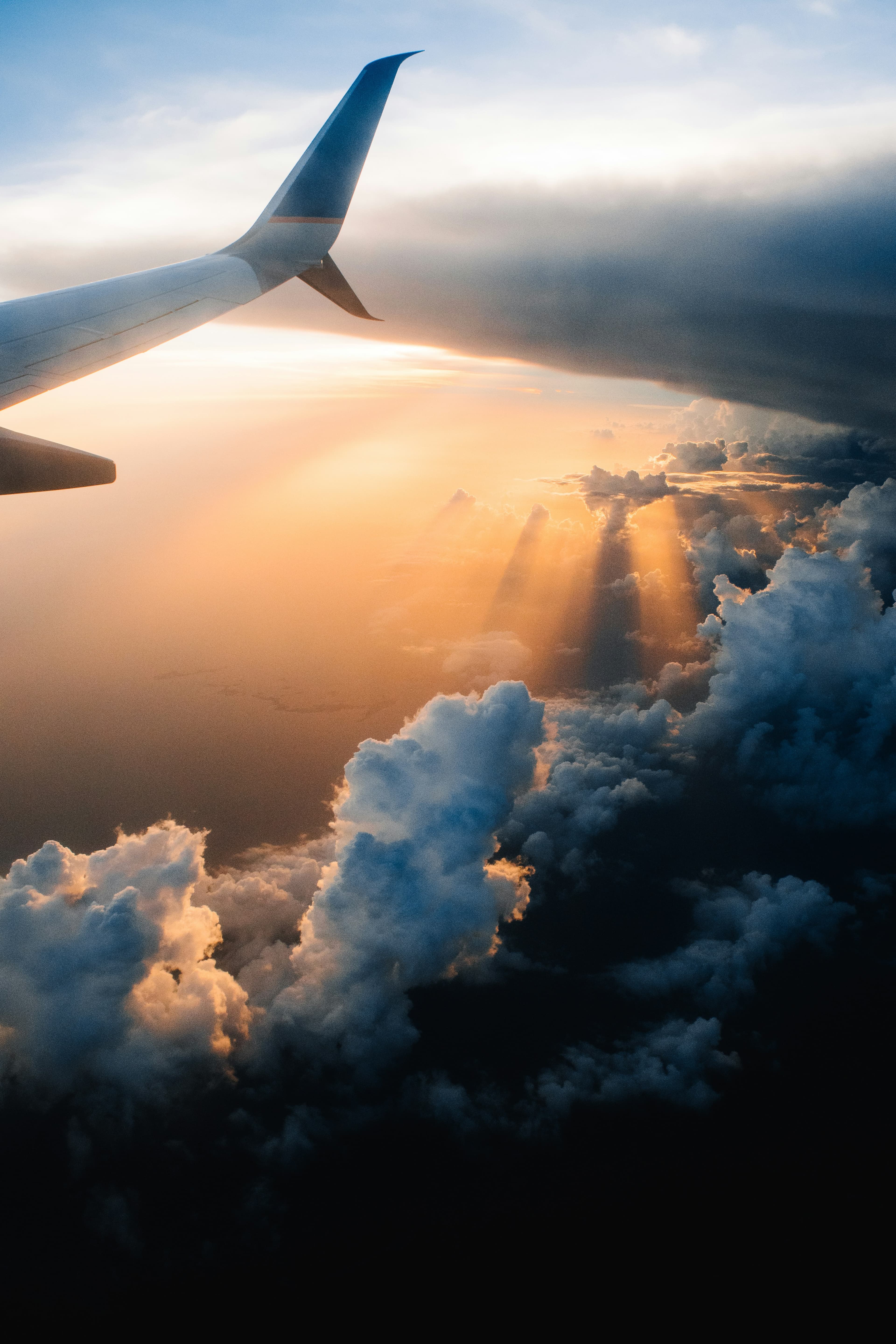 The wing of an airplane in the sky, above the clouds with golden rays of the sun shining through.