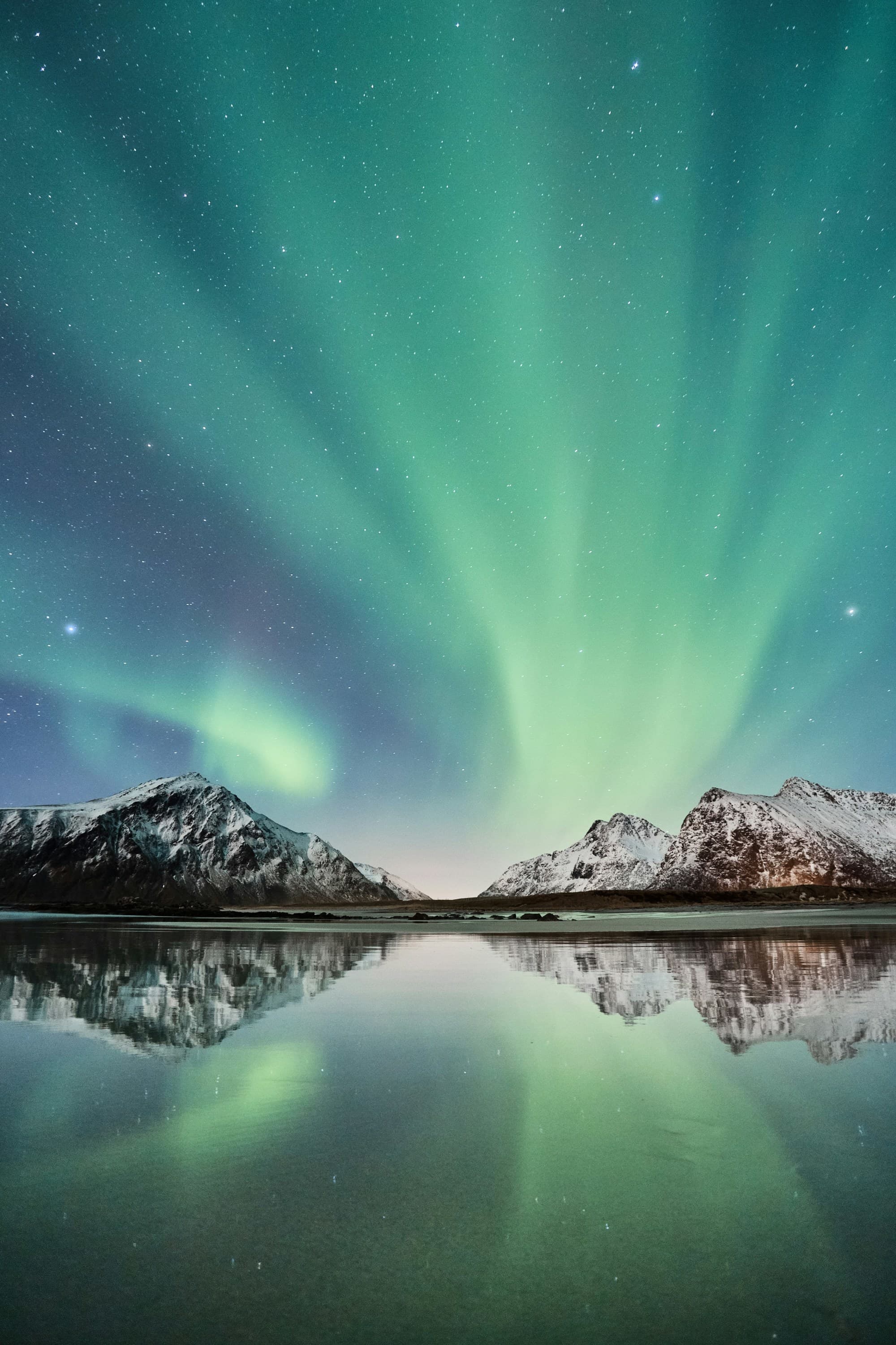 A stunning site of the Northern Lights and stars reflecting off of a body of water, with snow-covered mountains in the background.