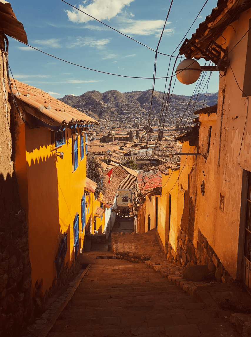 A vibrant view of yellow buildings with terracotta roofing and steps leading down to a view of the city with mountains in the far distance.