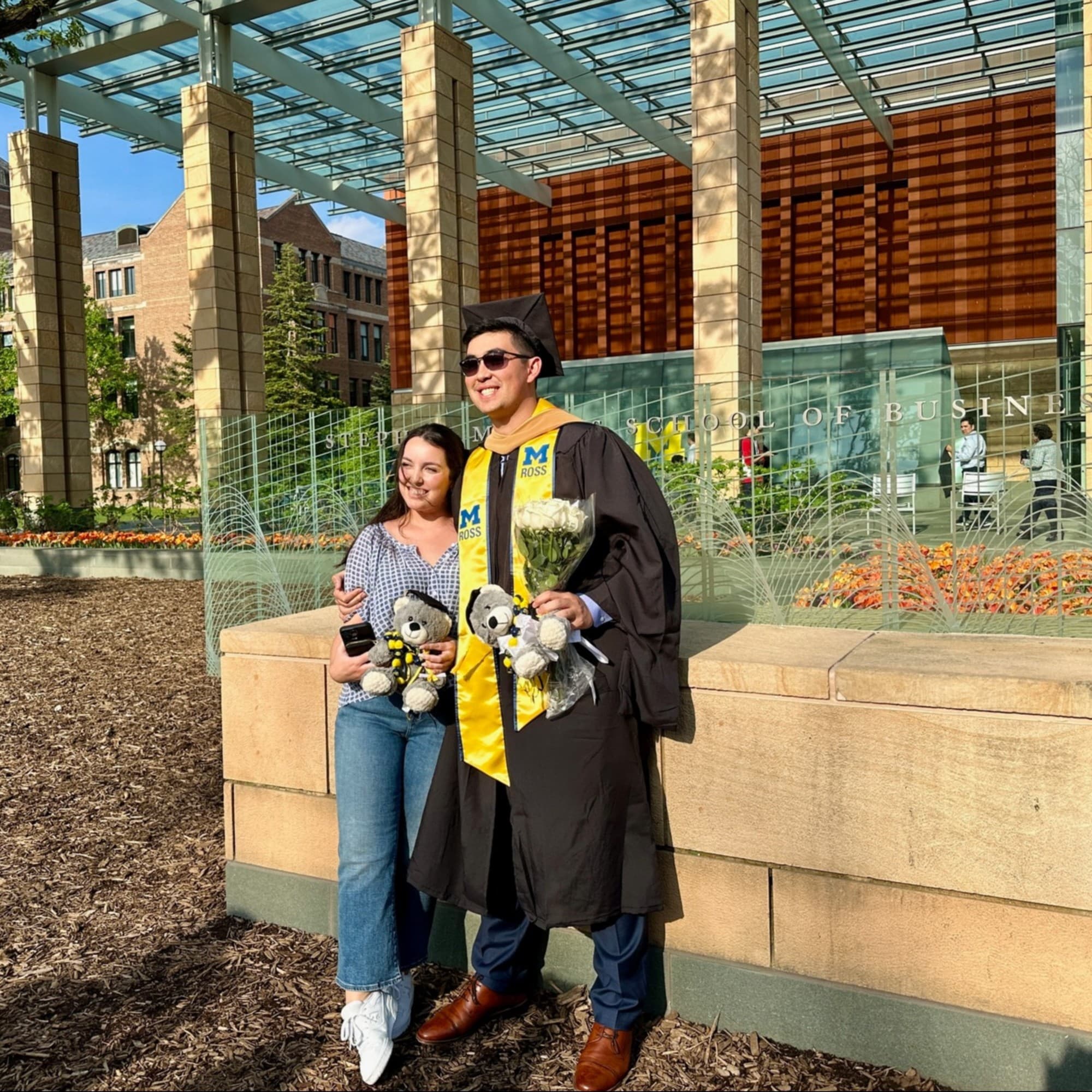 A graduate in cap and gown posing with another person for a photograph outside during the daytime