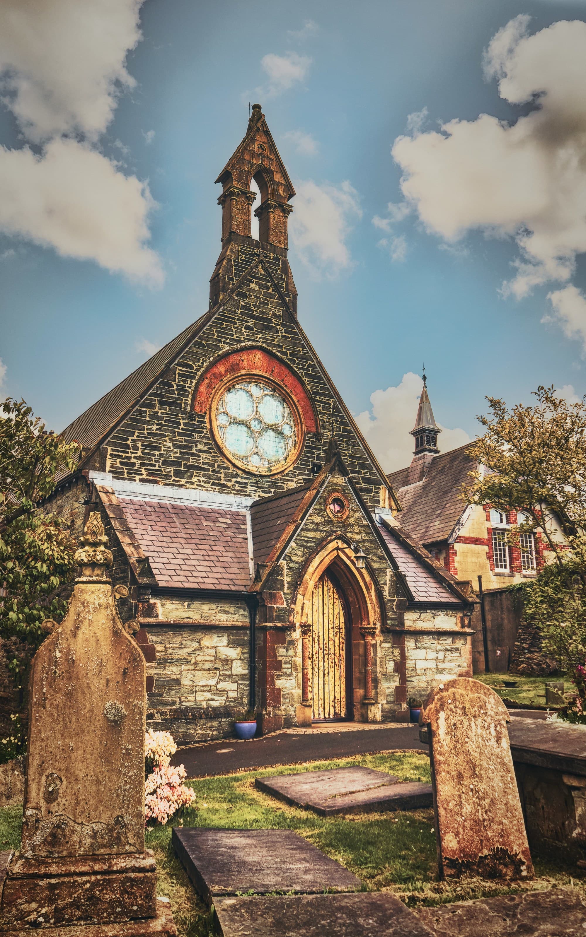 An old ornate building with a large circular window and a pointed spire, surrounded by gravestones under a blue sky with clouds.