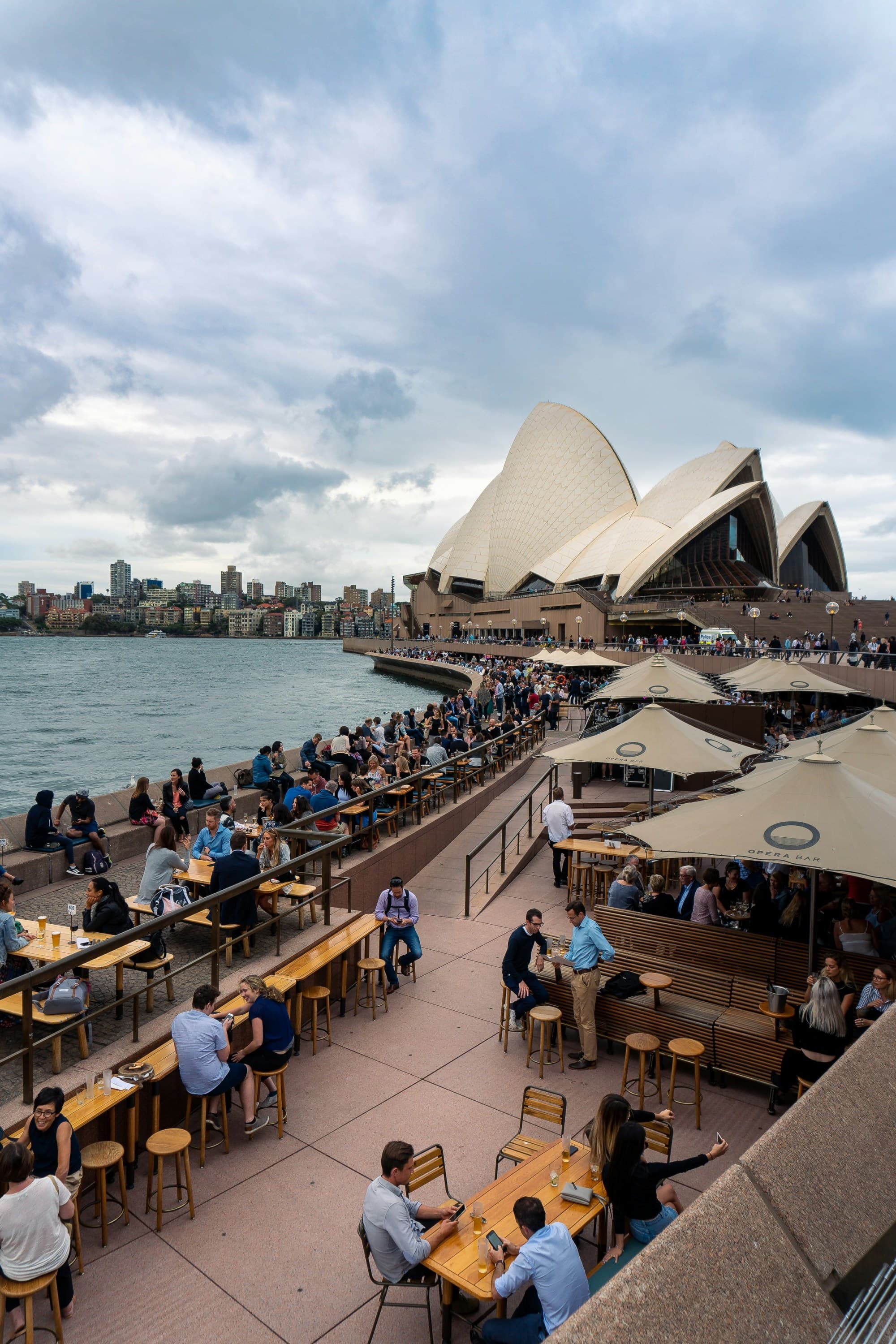 The image depicts an outdoor dining scene with guests enjoying a meal against the backdrop of the Sydney Opera House under a cloudy sky.