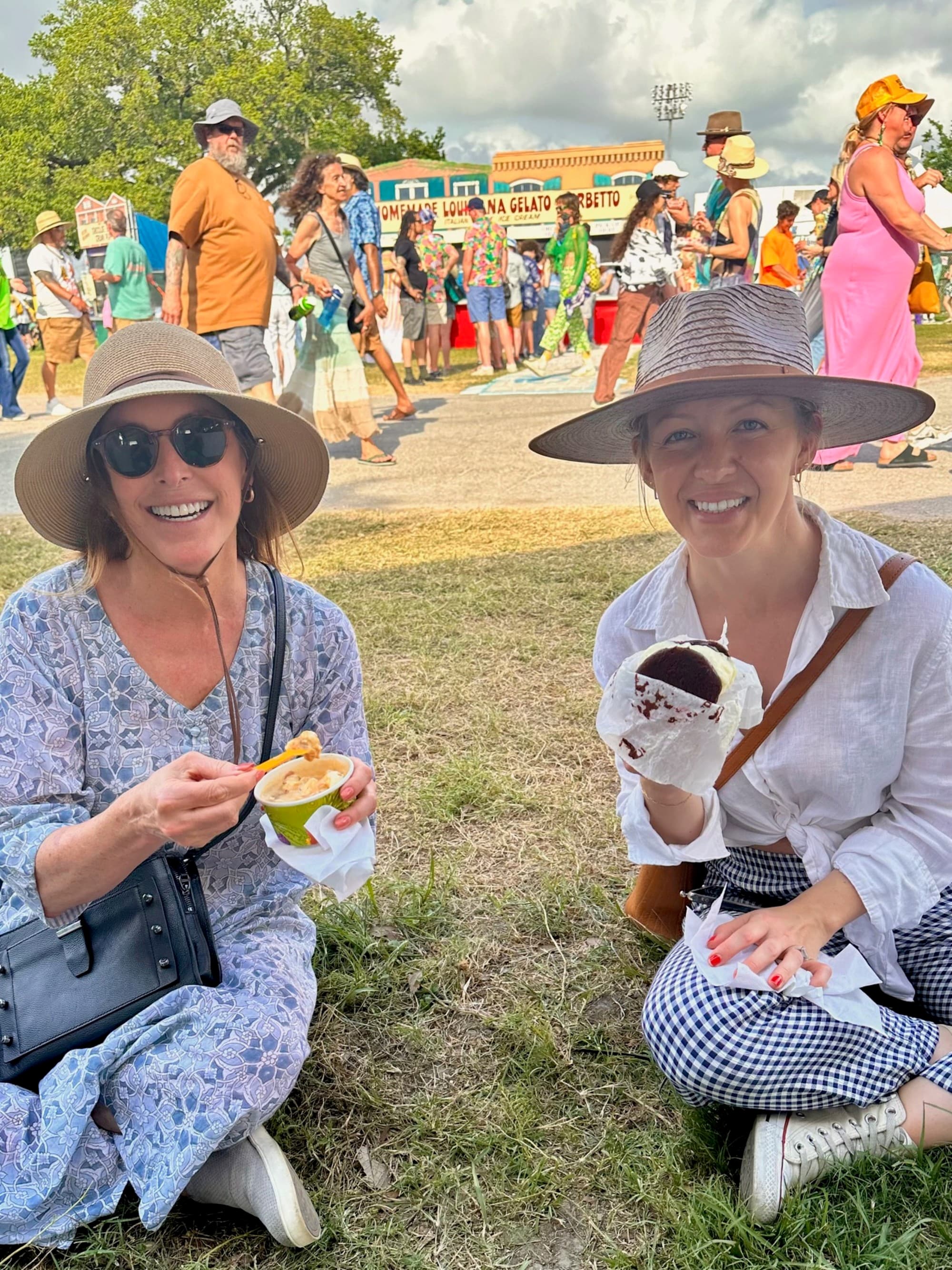 Allison and a friend in straw hats sitting on a lawn eating ice cream, with a lively crowd and food stalls in the background.