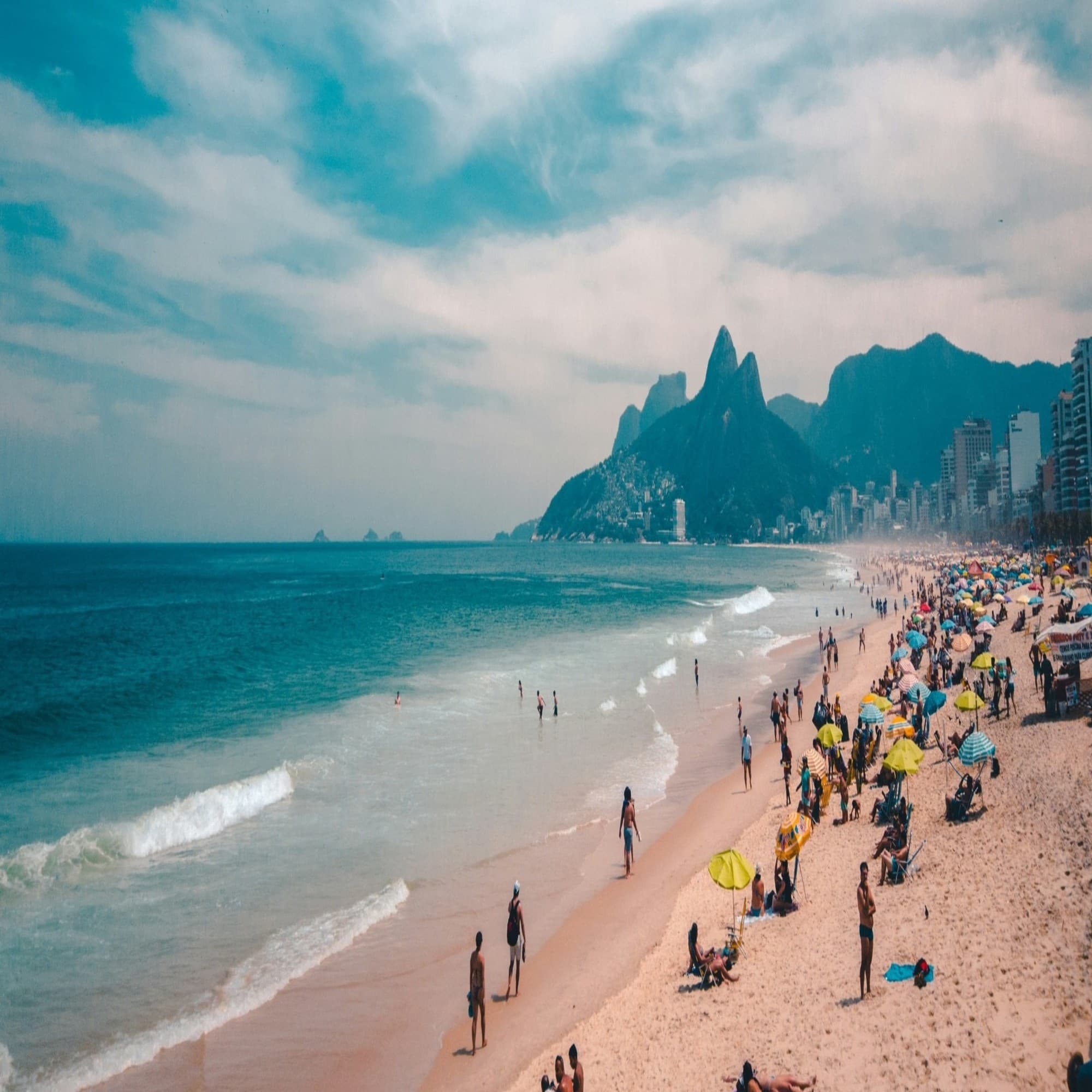 The image depicts a lively beach setting with a mountainous landscape in the background under a clear blue sky.