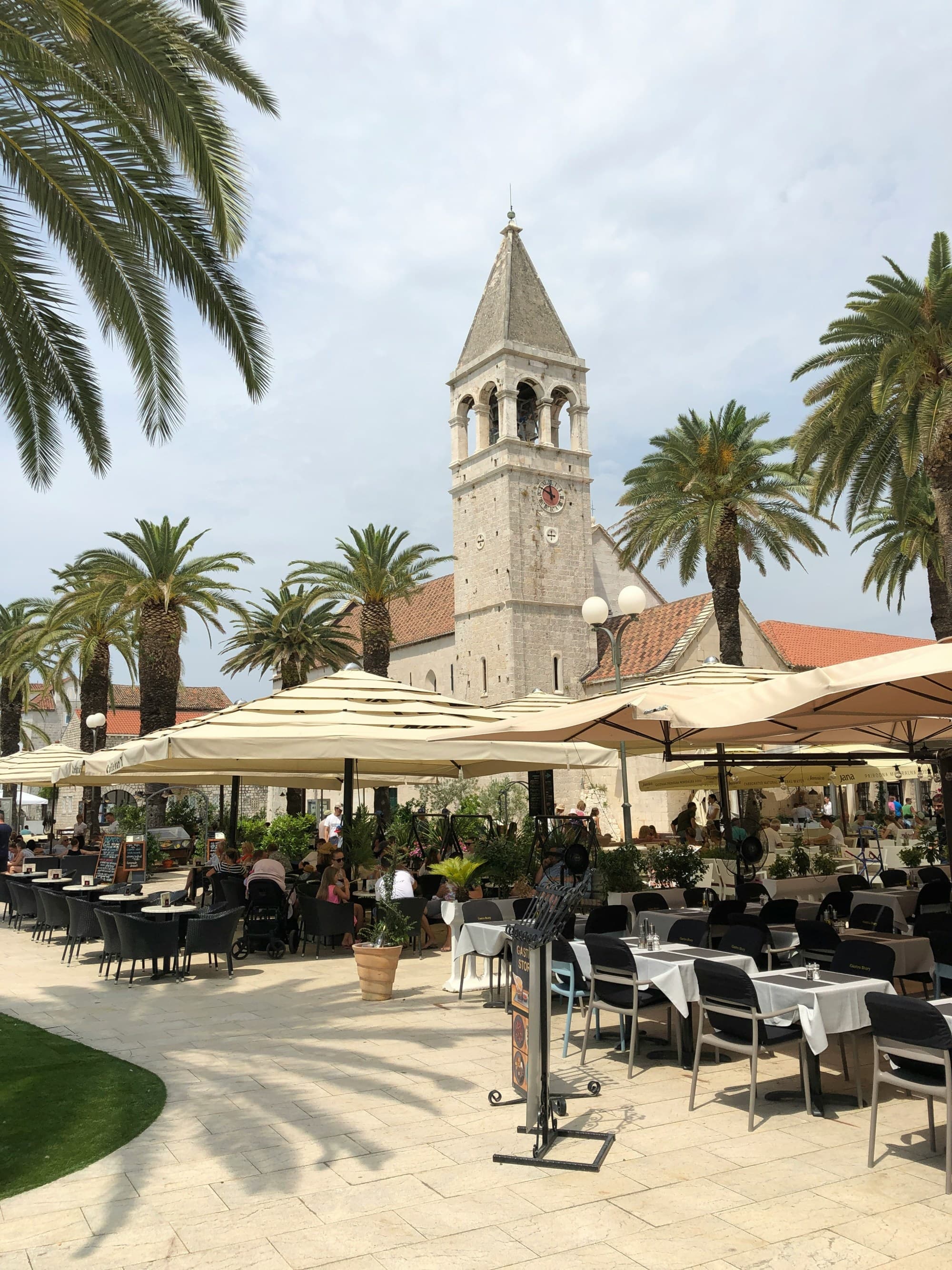 A bustling outdoor café scene with diners under umbrellas, flanked by palm trees and a historic bell tower in the background.