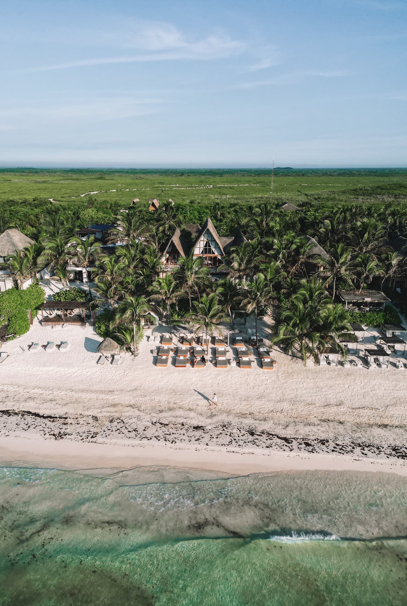 This image depicts an aerial view of a beach, ocean and villas aligned in row on the sandy shore. There is one large a-frame building in the background surrounded by lush green trees and foliage.