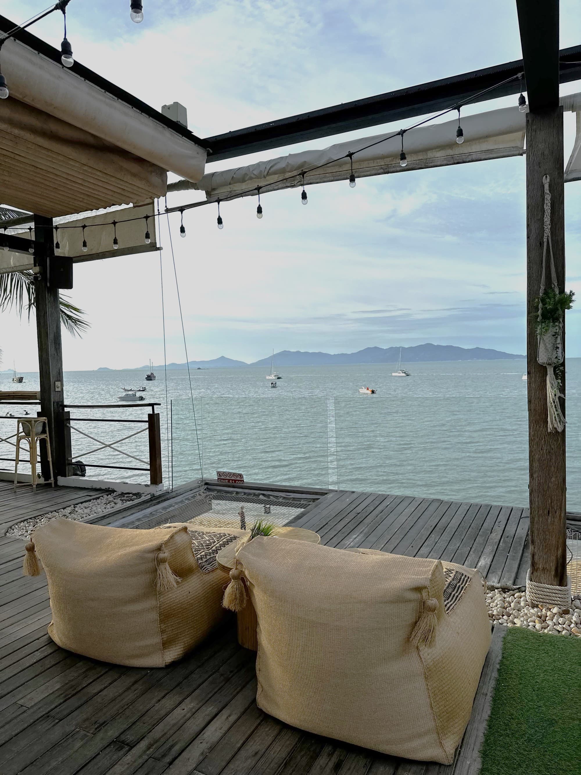 A serene seaside deck offers a relaxing spot with bean bag chairs and a view of boats under a cloudy sky.