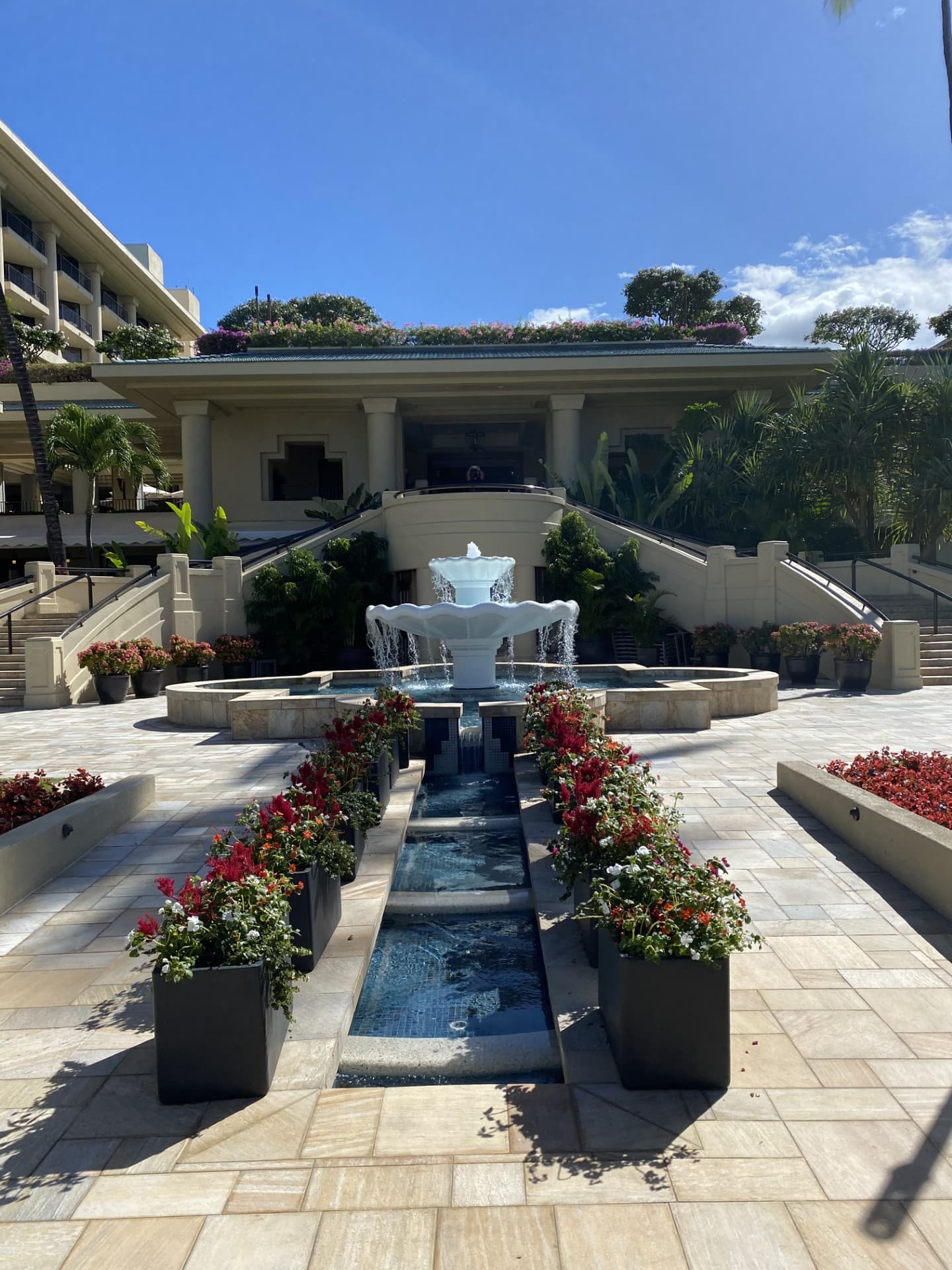 An outdoor fountain above a narrow, rectangular pool of water, bordered by red flowers, under a sunny sky.