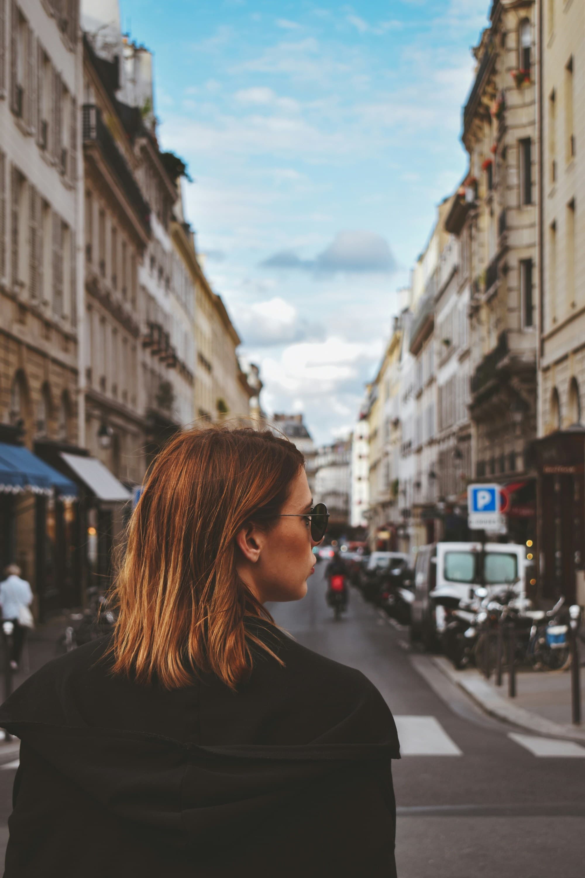 The image shows a city street scene with a person’s face blurred, amidst Parisian buildings and light traffic.