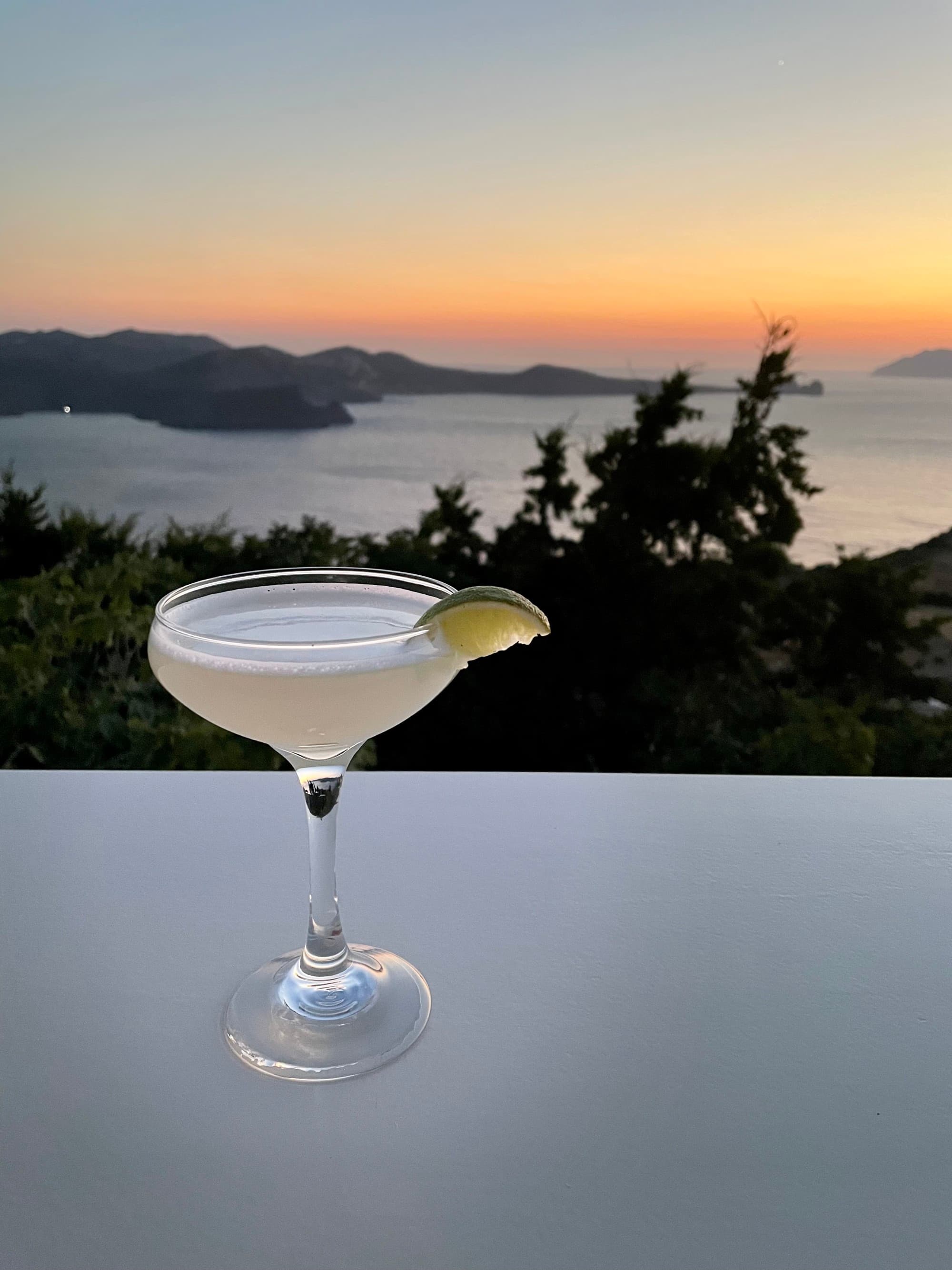 A close-up of an elegant cocktail set on a table, with a view of the sunset over the sea in the background.