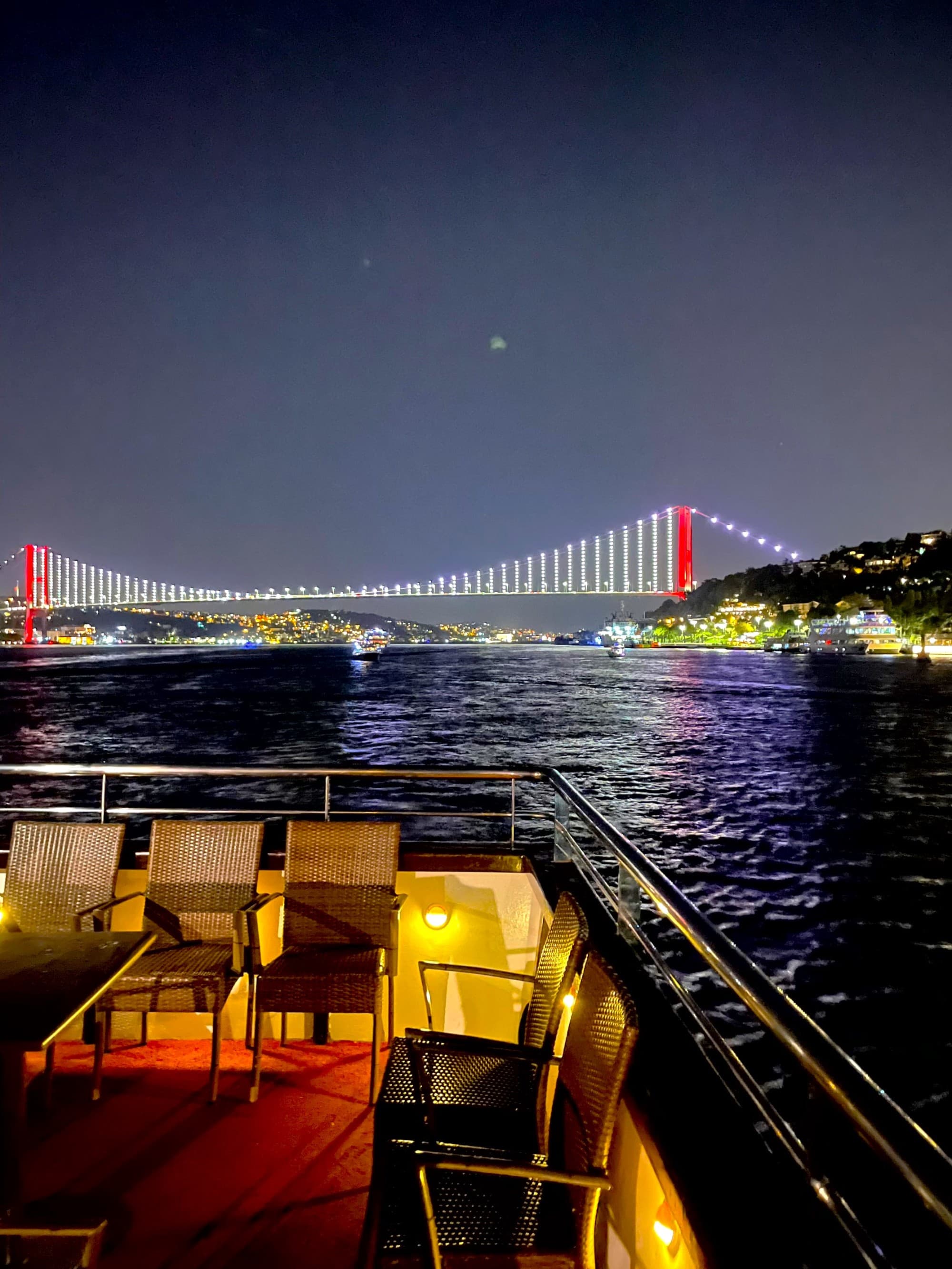 A beautiful view of chairs on a balcony overlooking a river and colorfully arched bridge in the distance at nighttime.