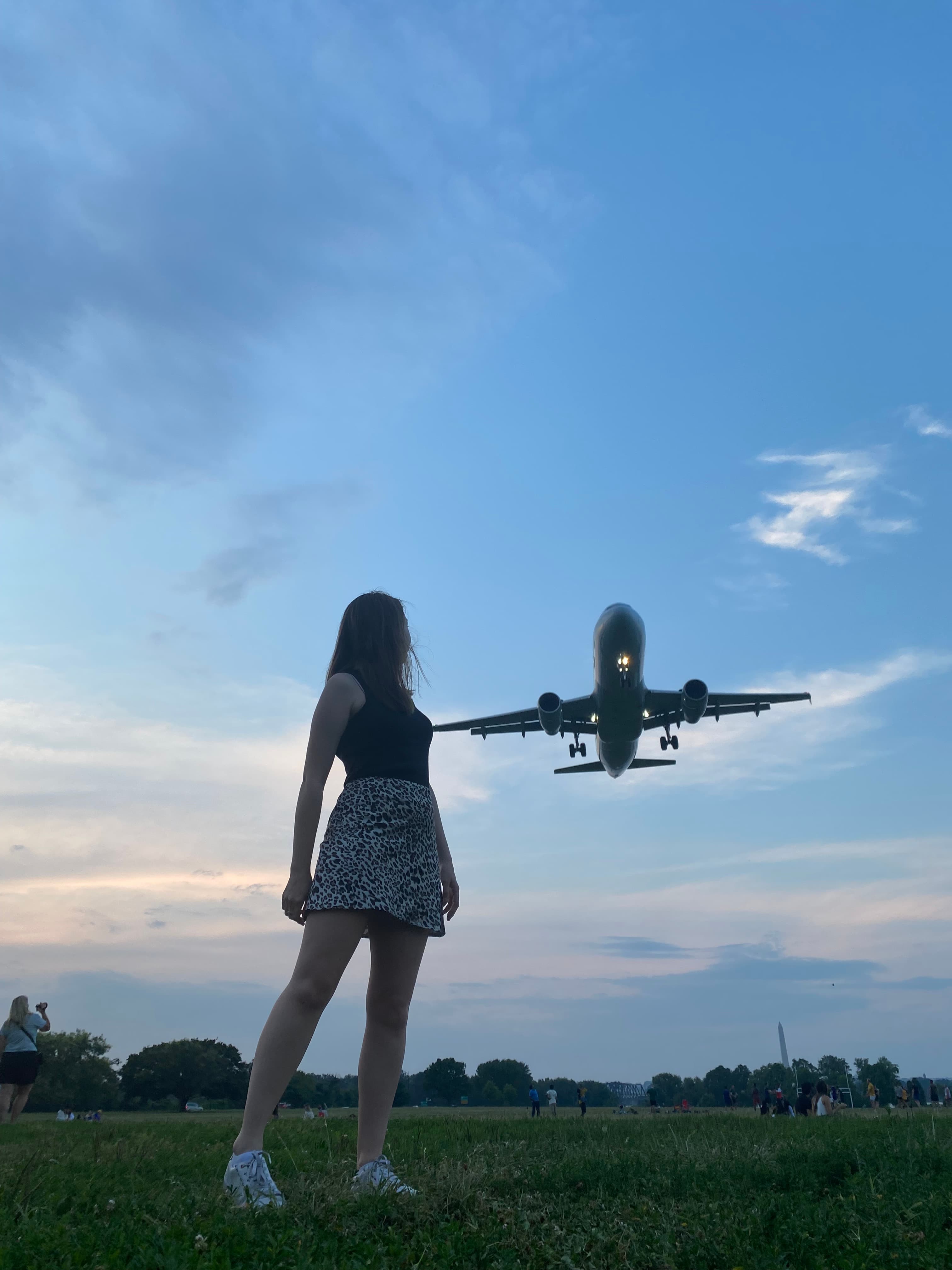 A woman standing on a grassy lawn outside and looking up to an airplane flying in the sky.