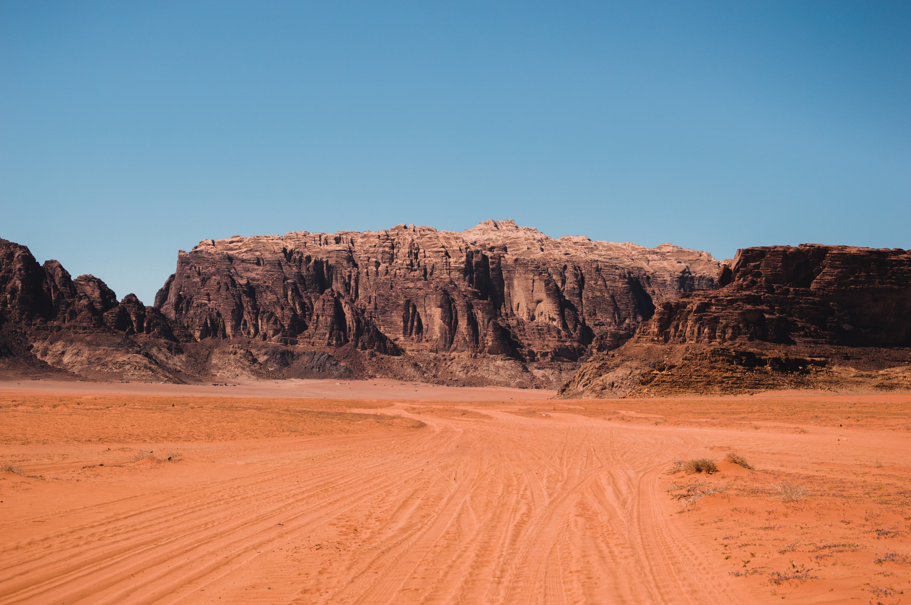 A visit to Jordan with red dirt roads and dark brown and red rock structures against a blue sky.