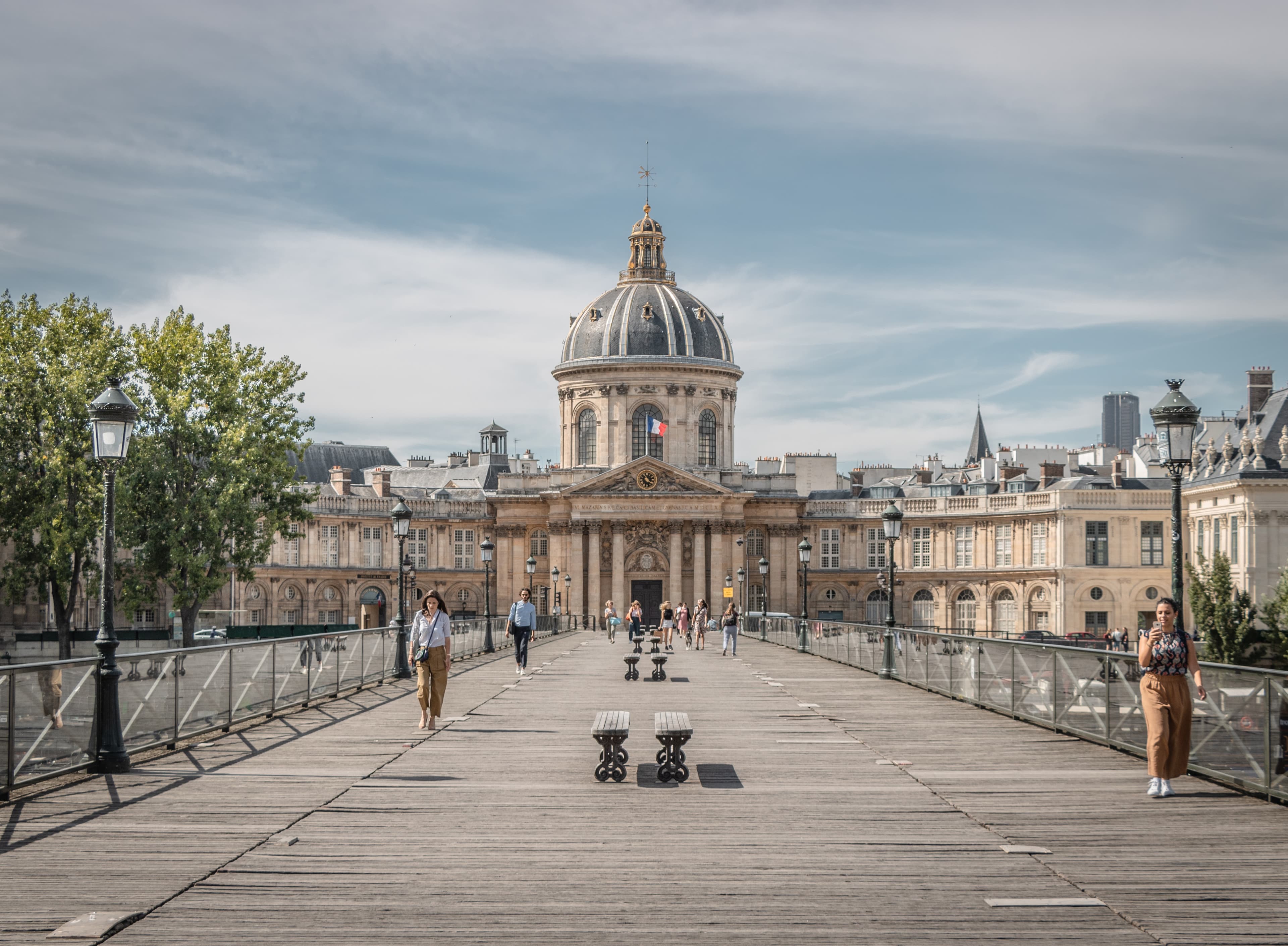 A brown bridge in Paris with people walking and a tan building with a blue rounded roof in the center with green trees