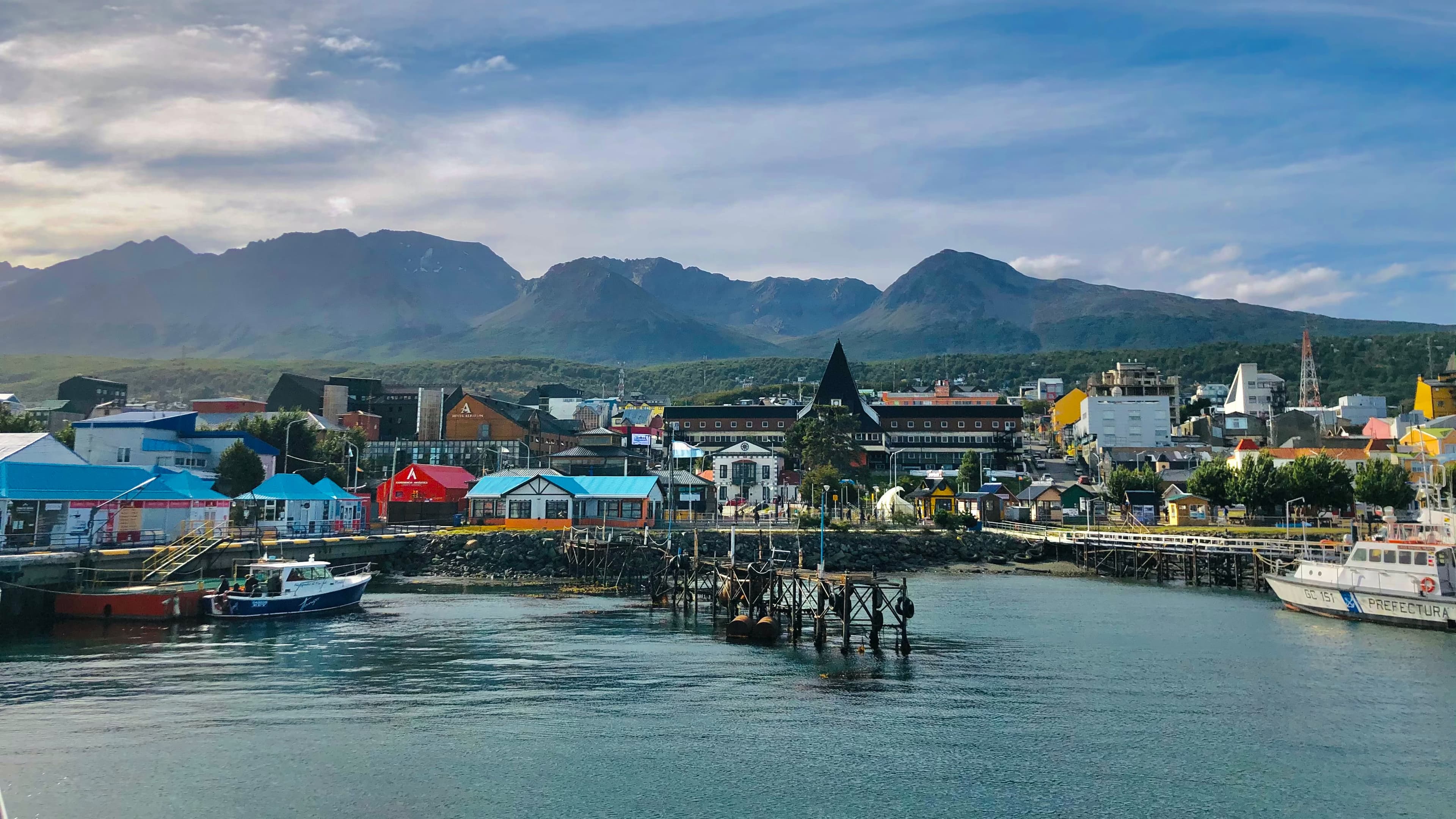 A blue lake with pink, blue and yellow houses and boats with green and brown mountains in the distance in Ushuaia on a Argentina vacation.