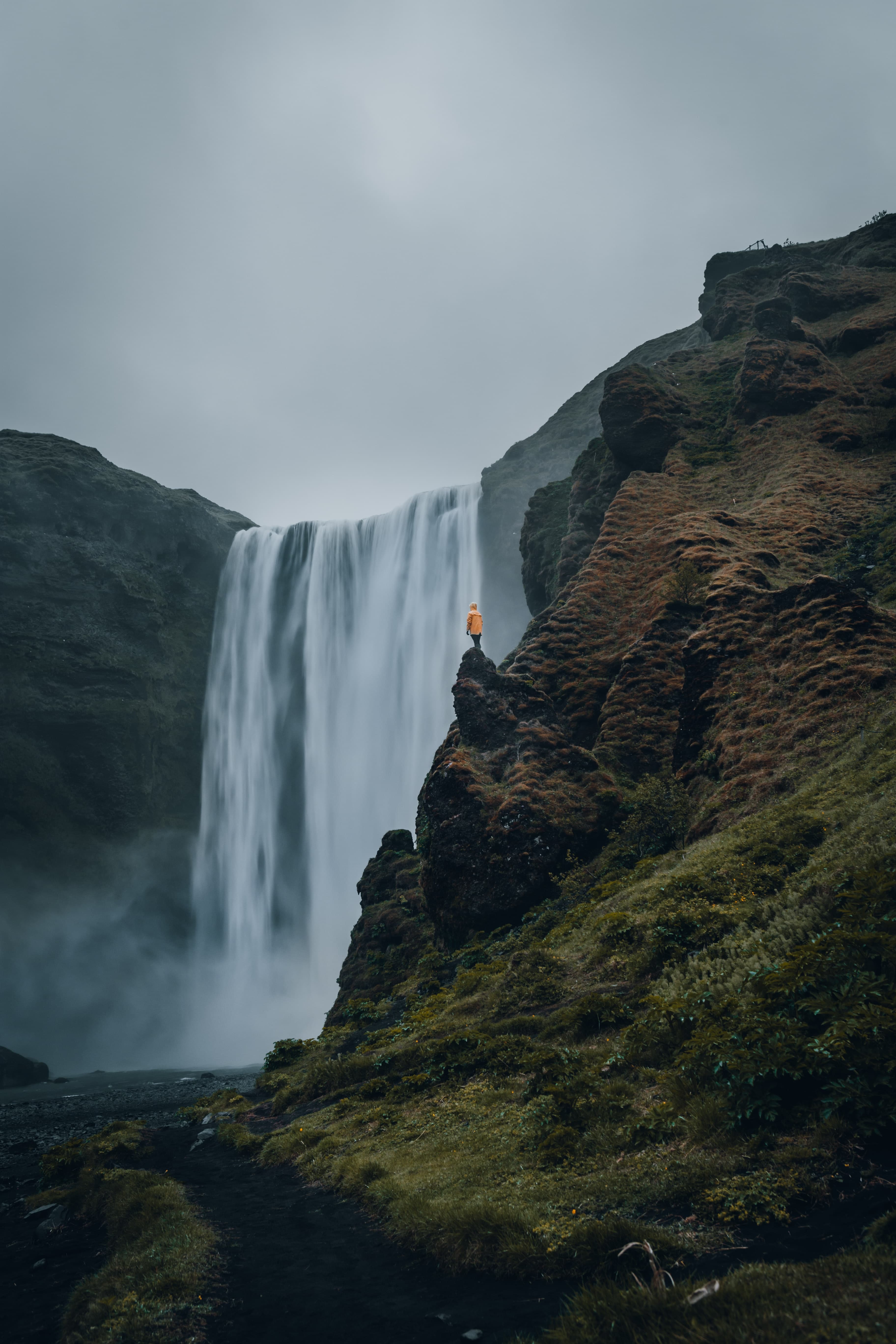 Person wearing orange jacket standing in front of waterfall on a cloudy day