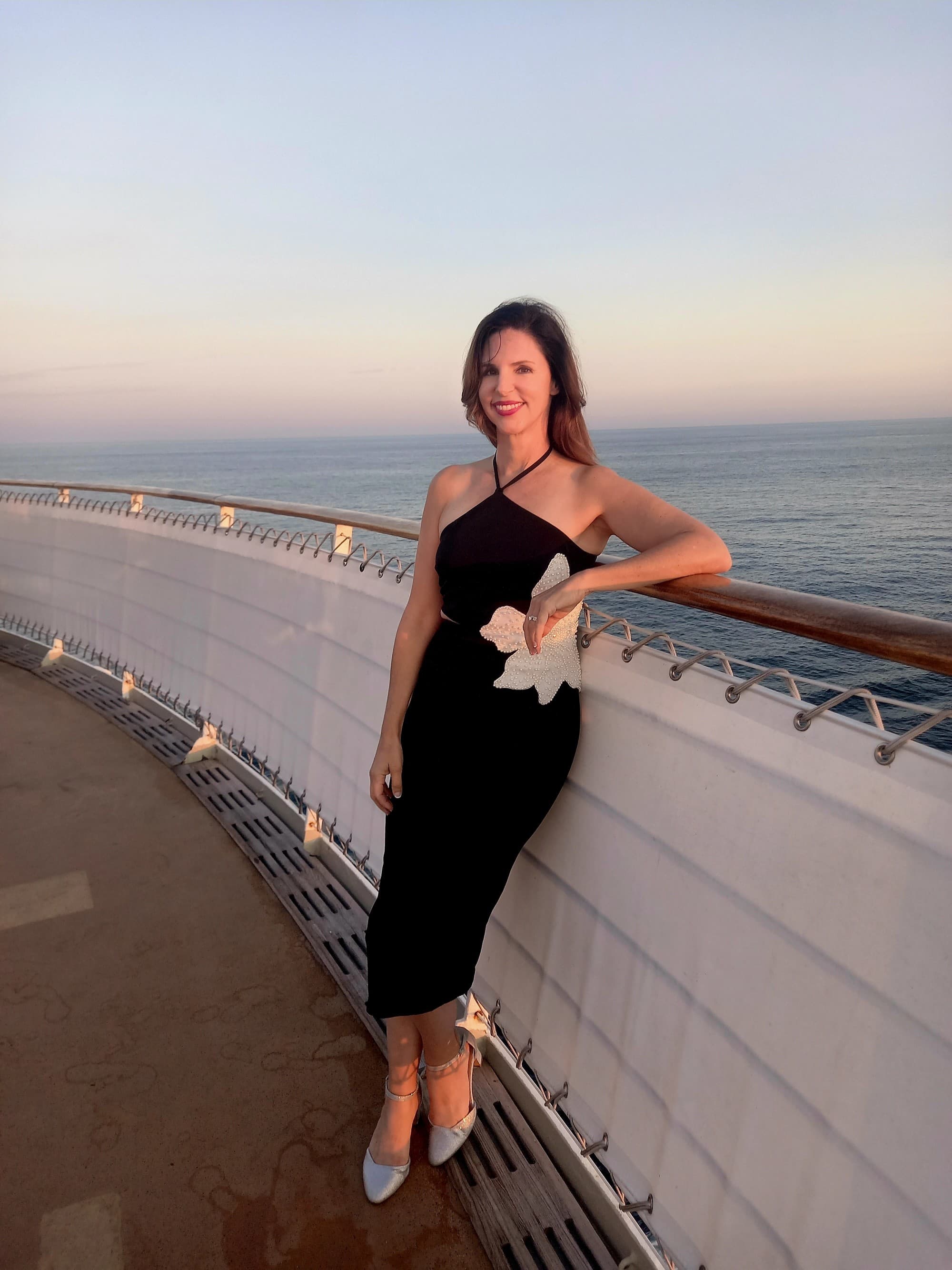 A woman in a black dress posing against the railing of a ship deck.