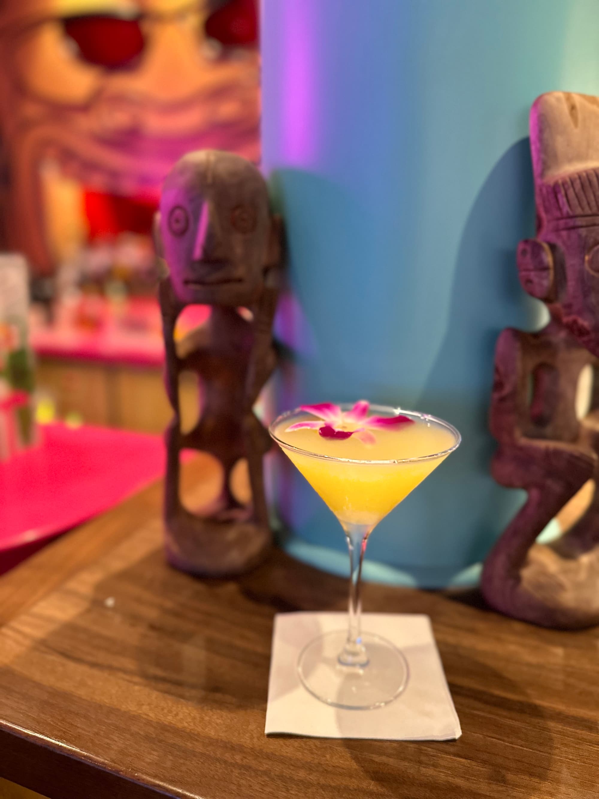 A yellow cocktail with a pink flower garnish sits on a wooden bar with tiki decorations around it at Pesky Parrot Tiki Bar