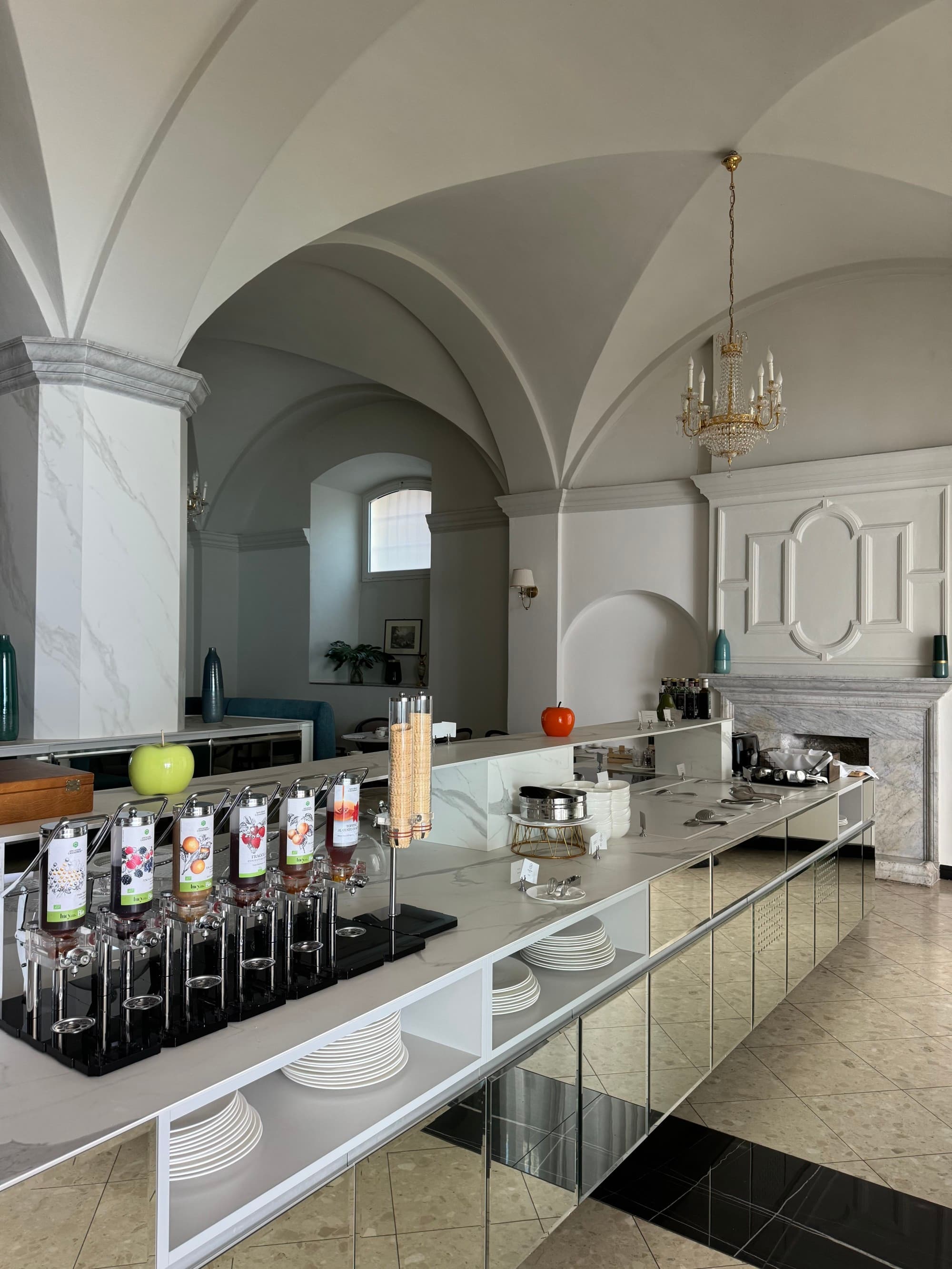 A view of a white room with tall arched ceilings and a chandelier hanging with an ice cream bar at Hotel Royal Victoria.