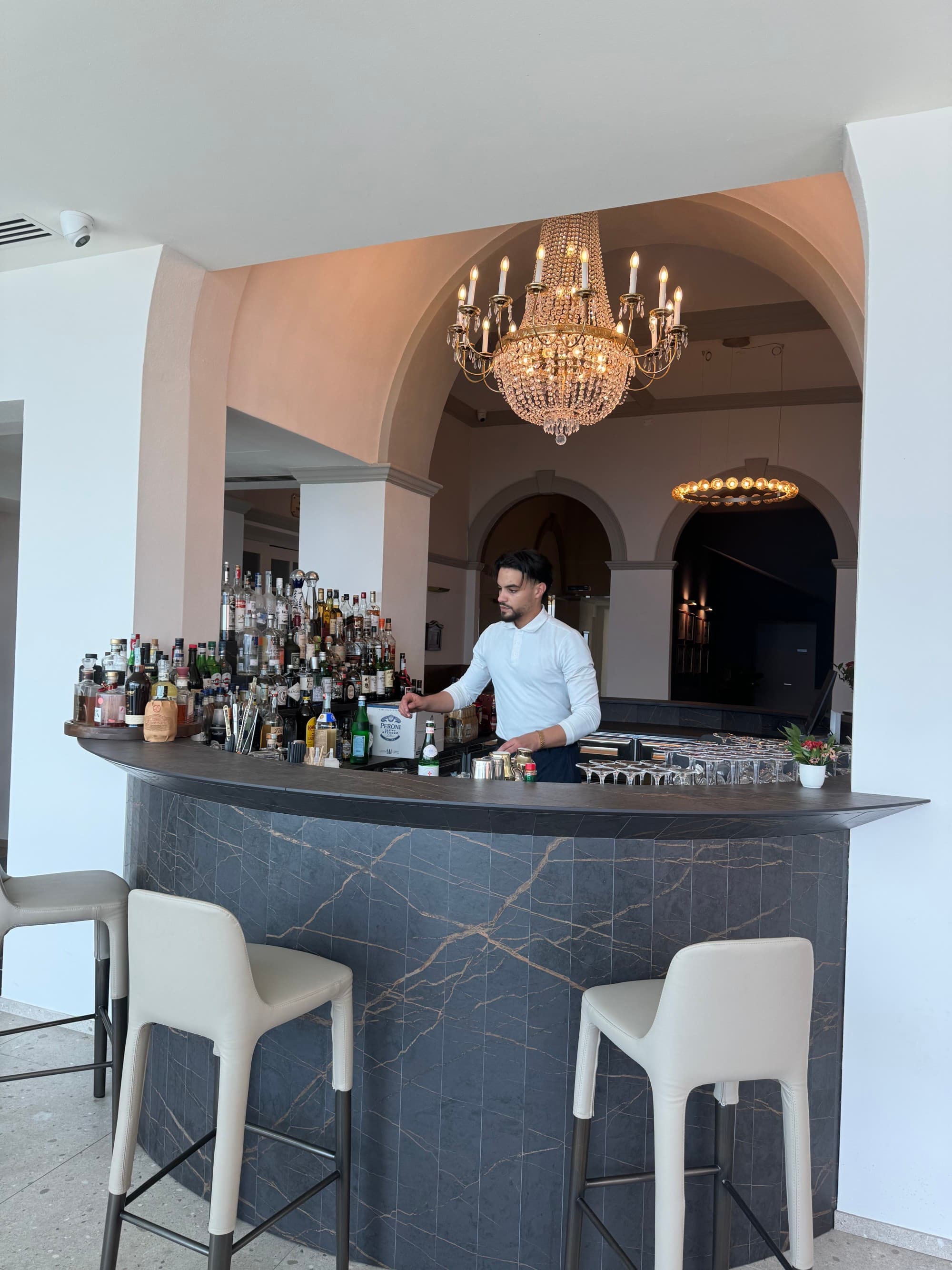 A bartender standing behind a black curved bar with a chandelier hanging above them at Hotel Royal Victoria Bar