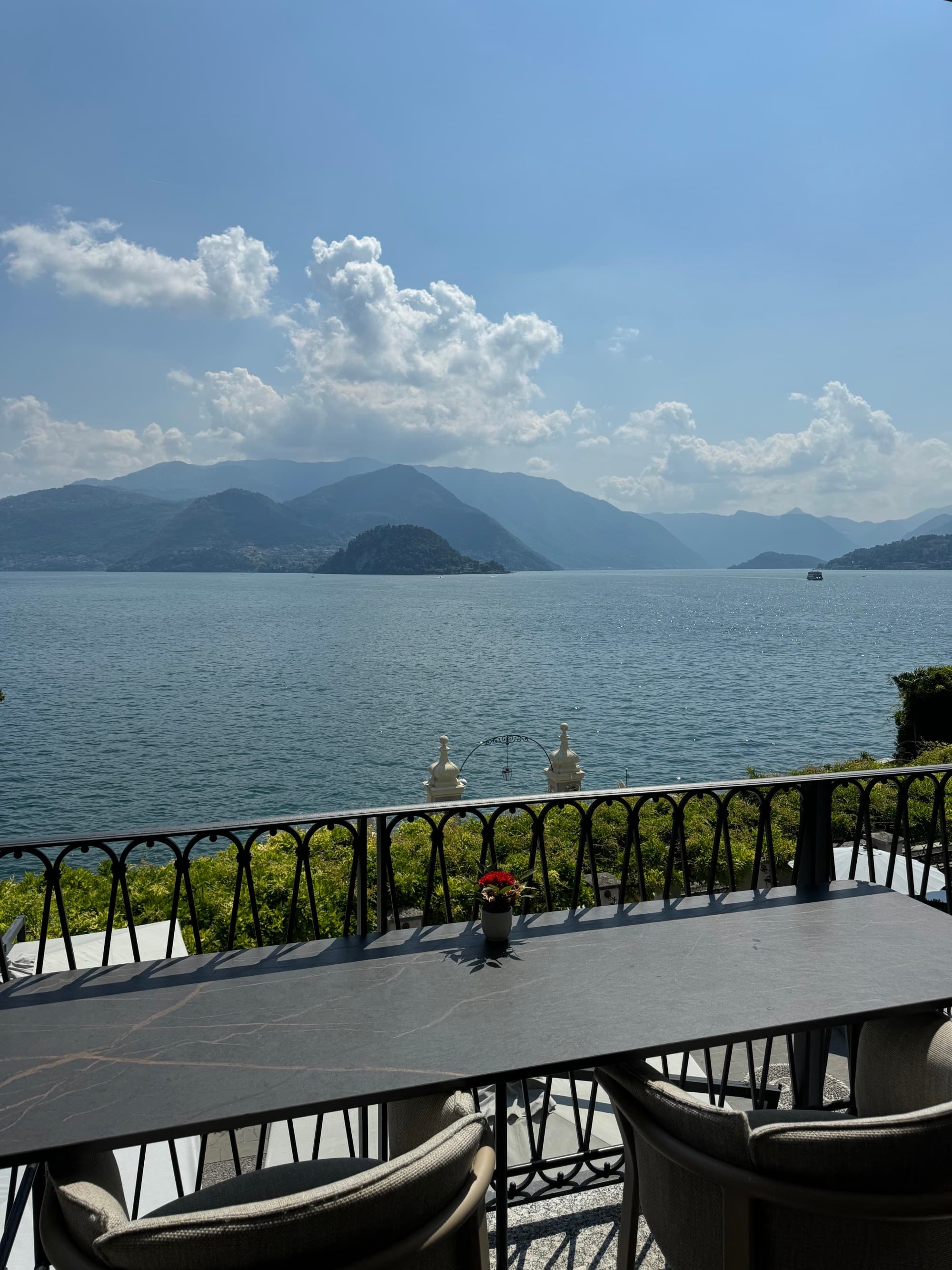 A lakeside view from a balcony, calm water, partly cloudy sky and mountains in the distance.