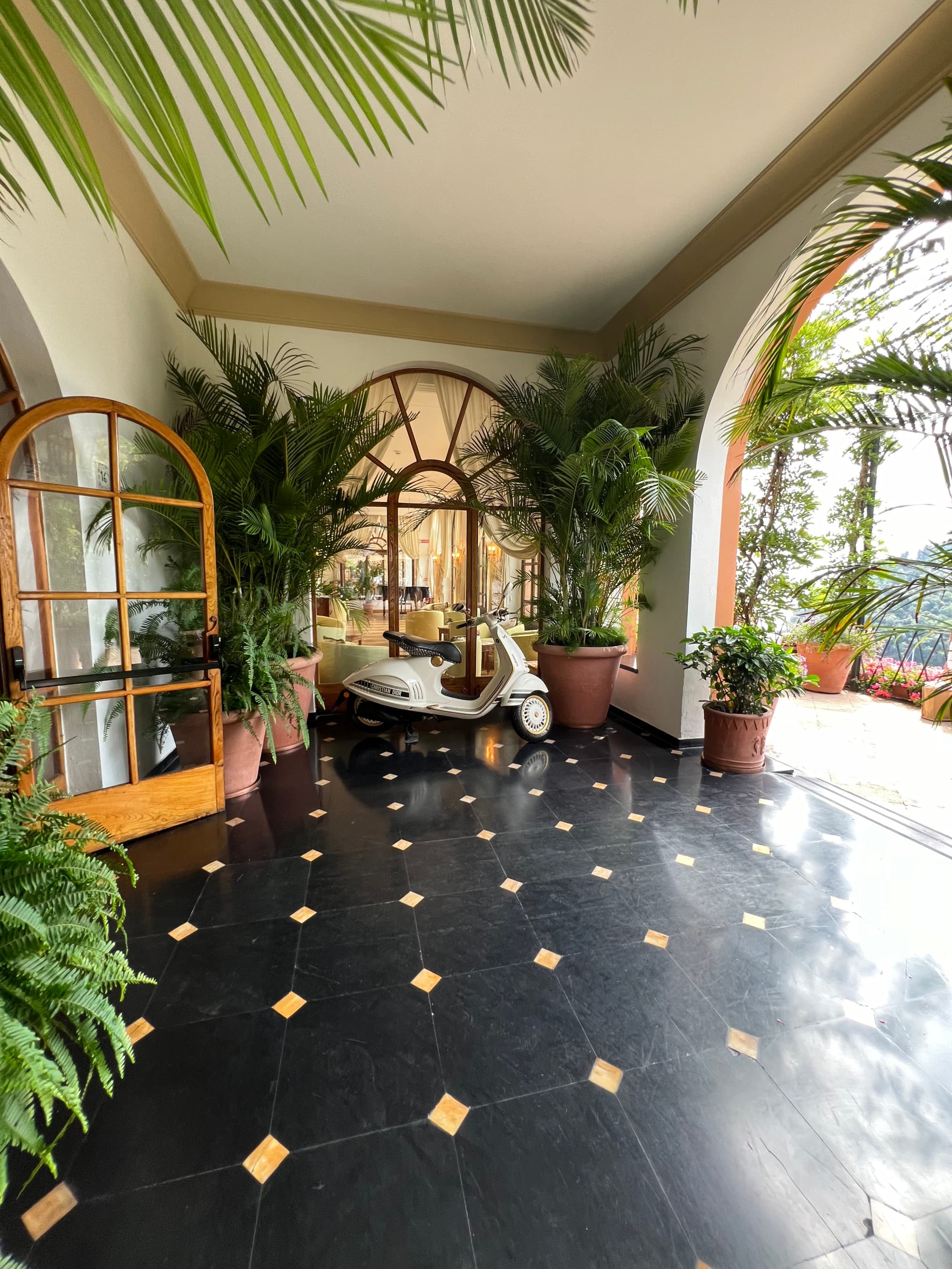 A view of the hallway, with black and gold tile floors, arched windows and doors and lots of potted palms lining the walls.