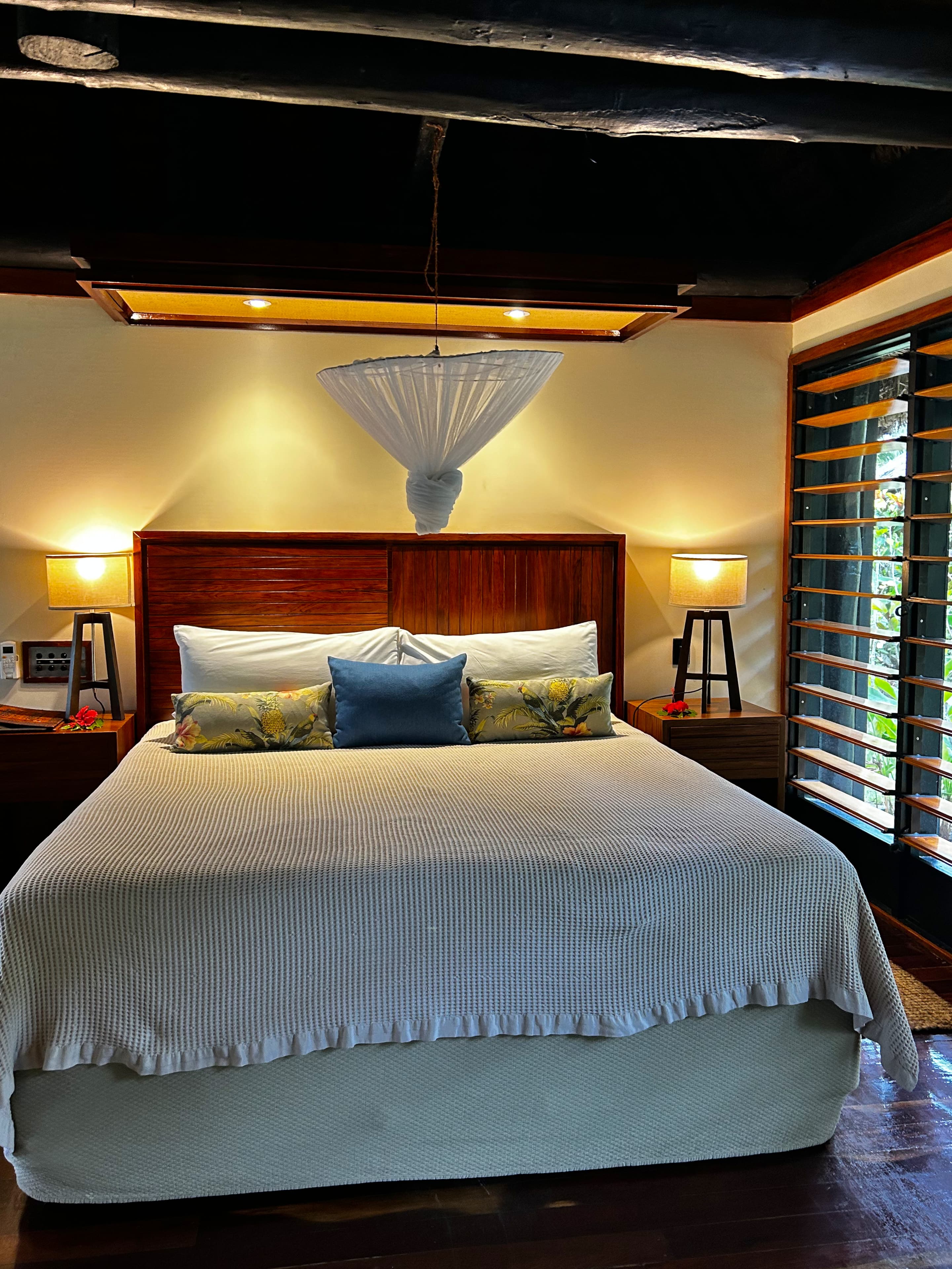 A view of the interior of a hotel room with a bed with wooden headboard and white mosquito tied above.