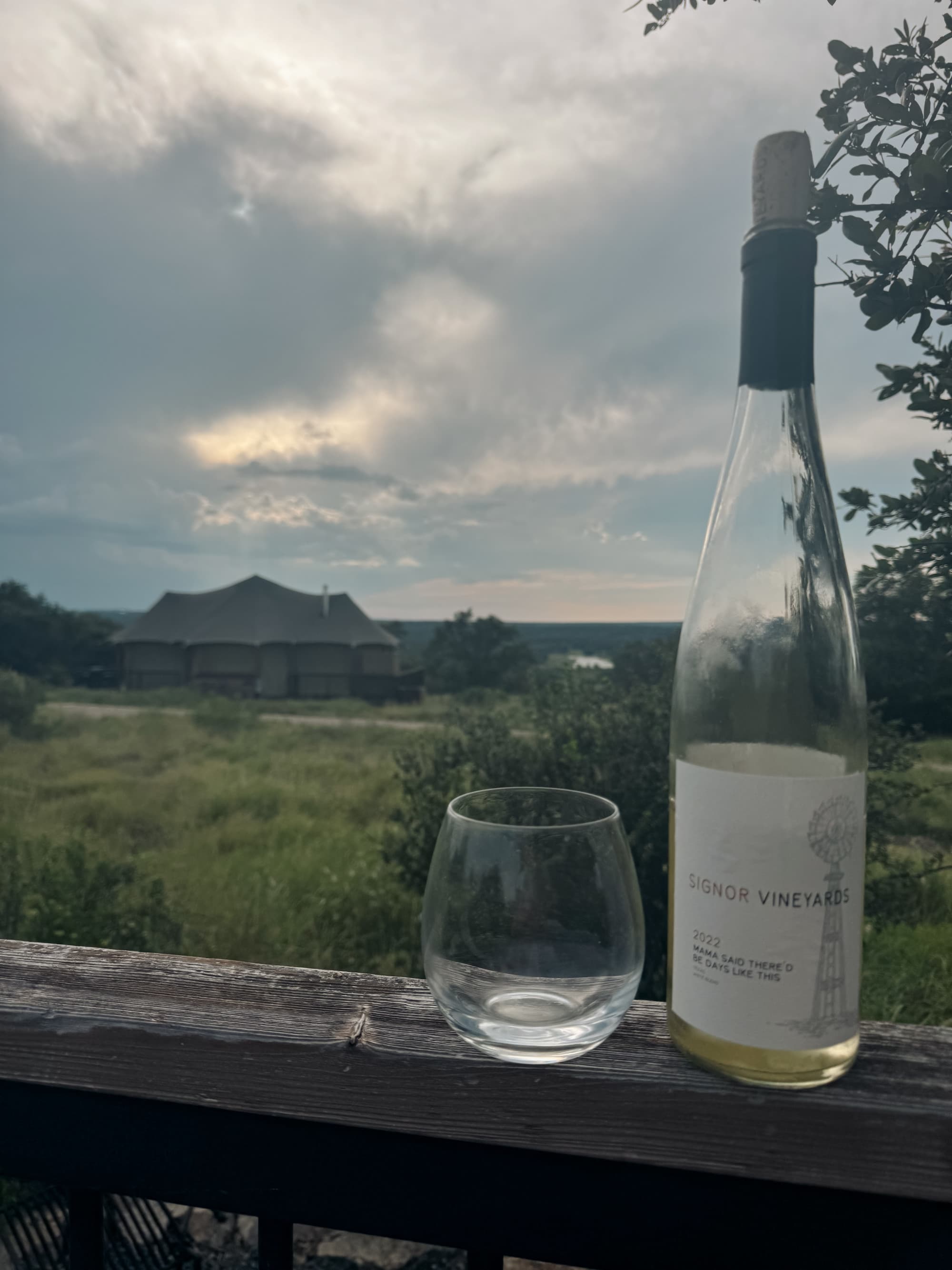 A bottle of wine and empty glass sitting on a railing with a view of a field