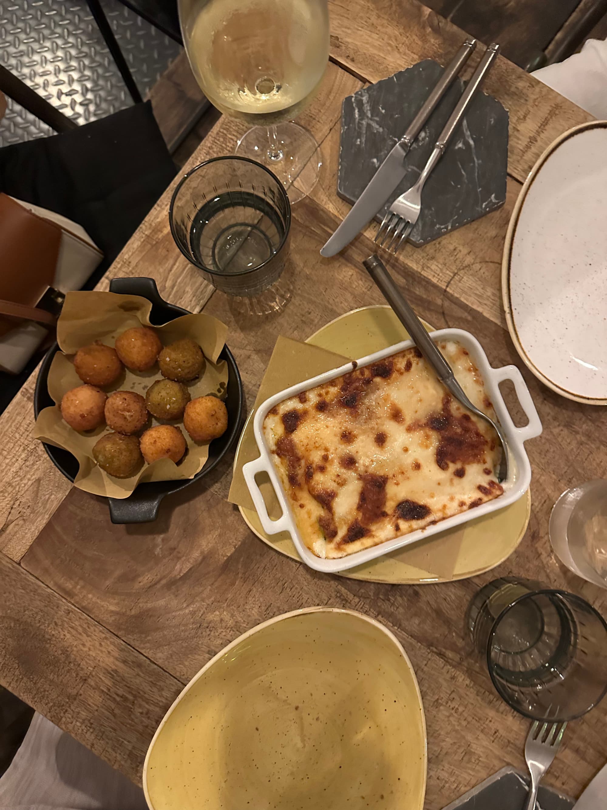 A food view of lasagna, bread and table settings on a wooden table at Osteria - Georgia Goddard