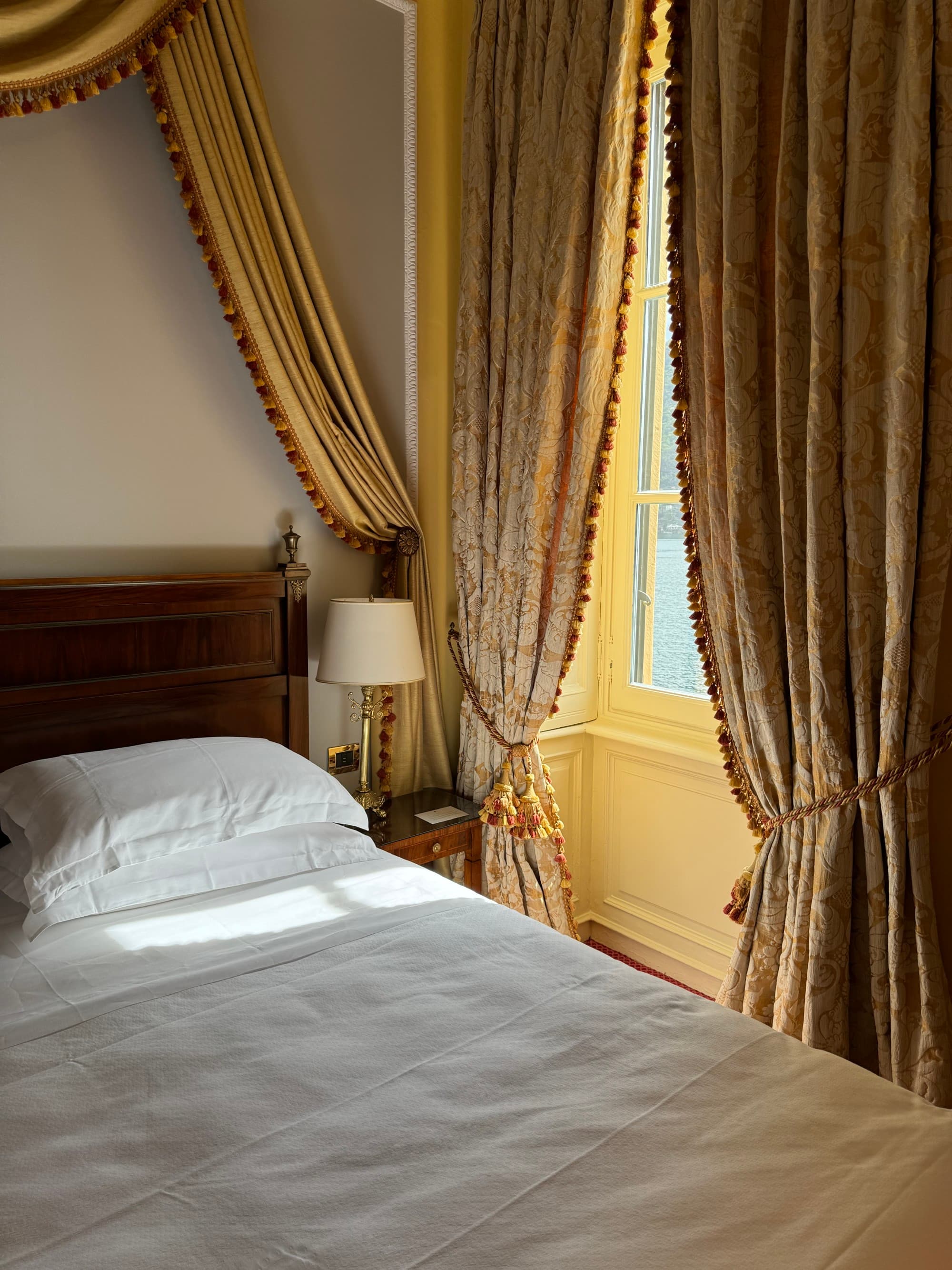 The hotel room view of the bed against a brightly-lit window covered with rich drapes.