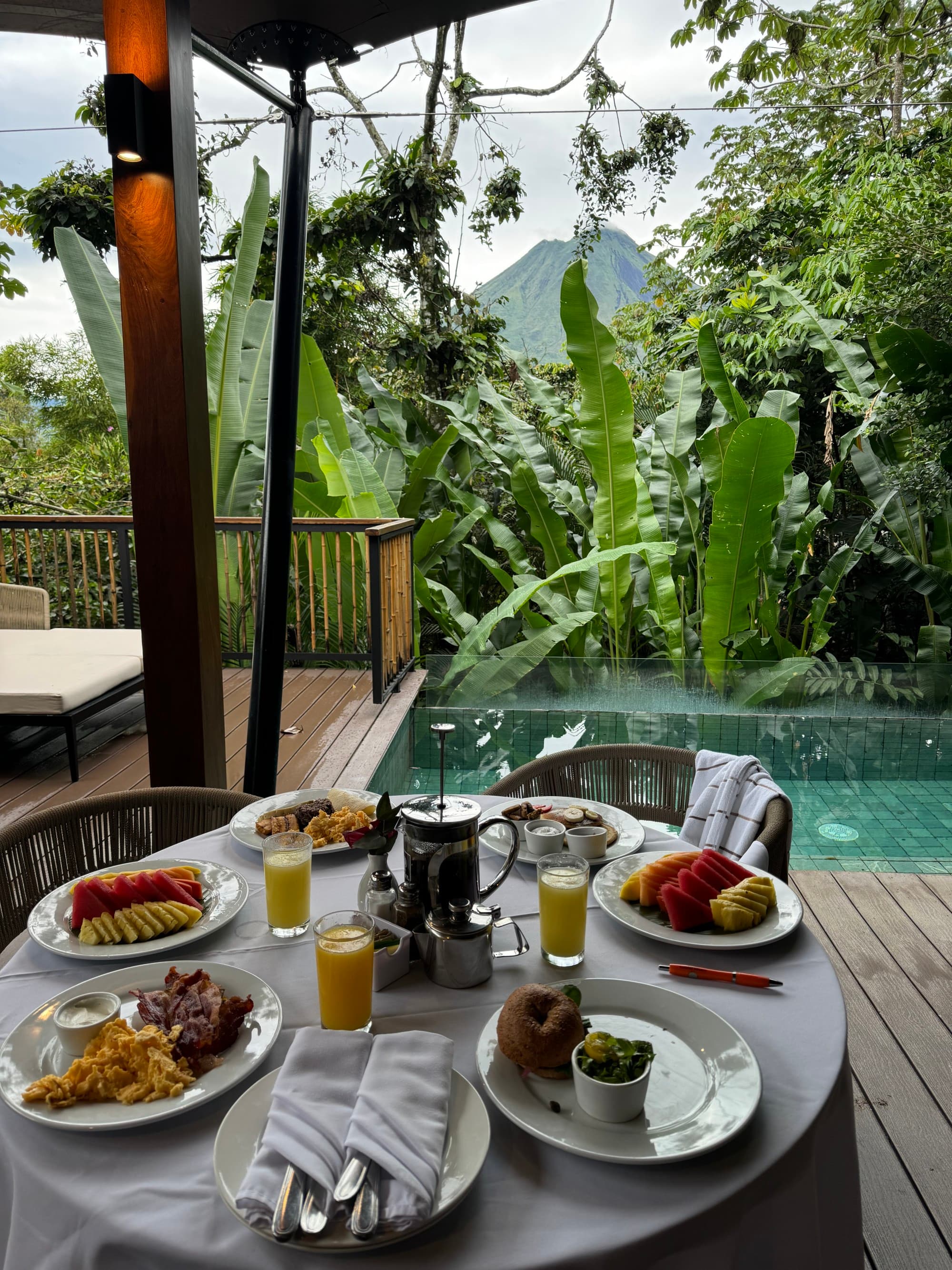 Decadent breakfast on a table next to a pool with a jungle in the background.