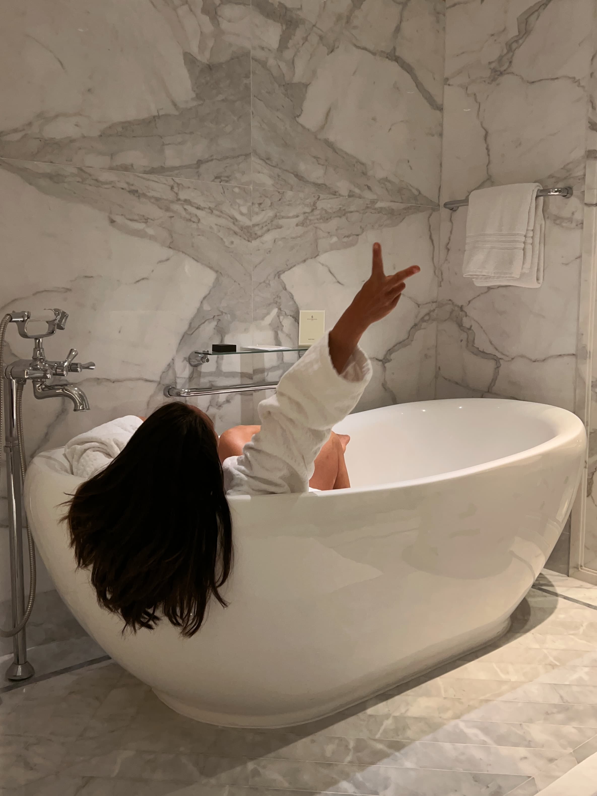 Travel advisor lying in the bath tub in a robe in the hotel bathroom posing with her hand in the air