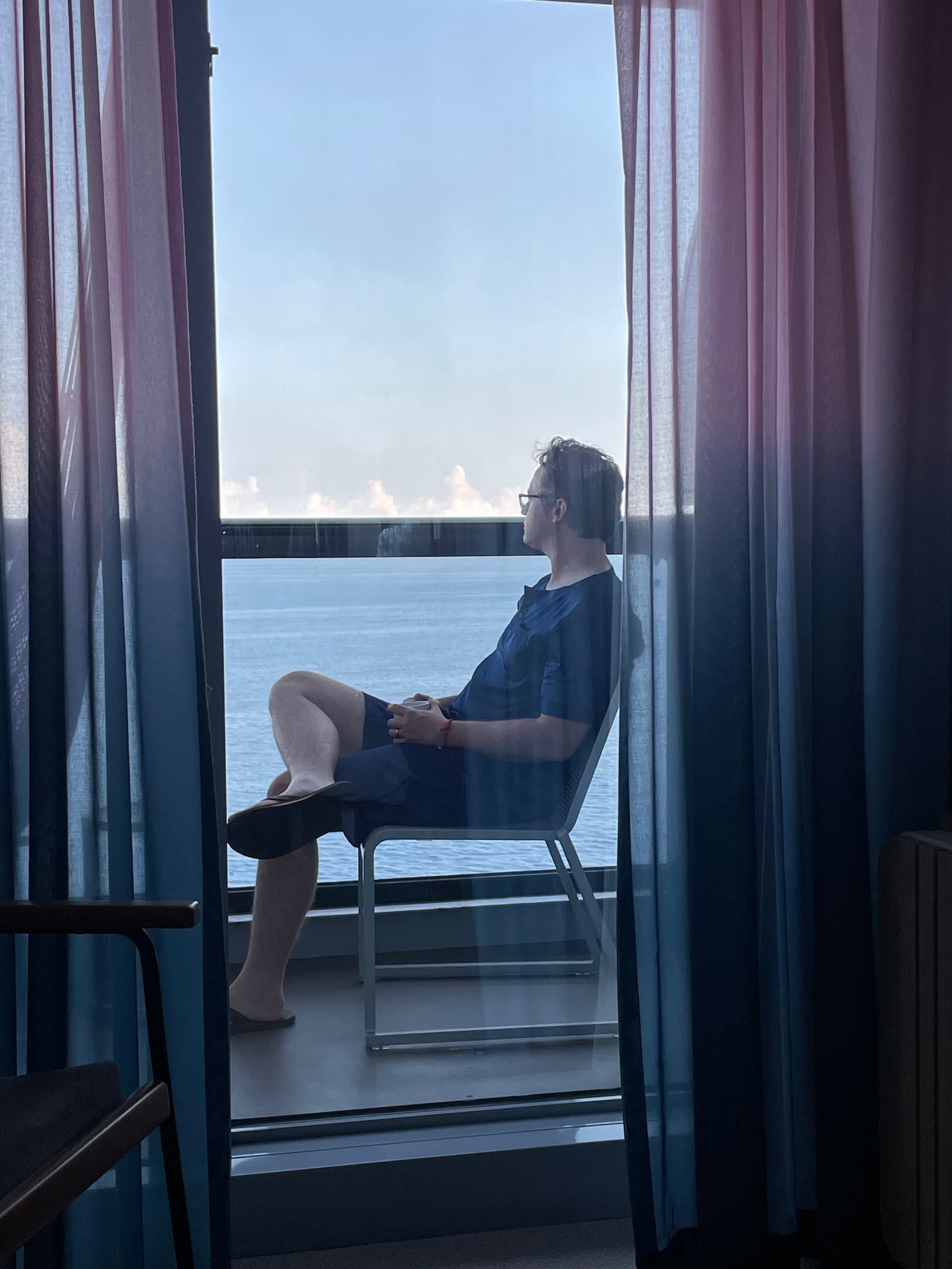 A man sitting on a chair on a balcony of a cruise ship admiring the ocean view.