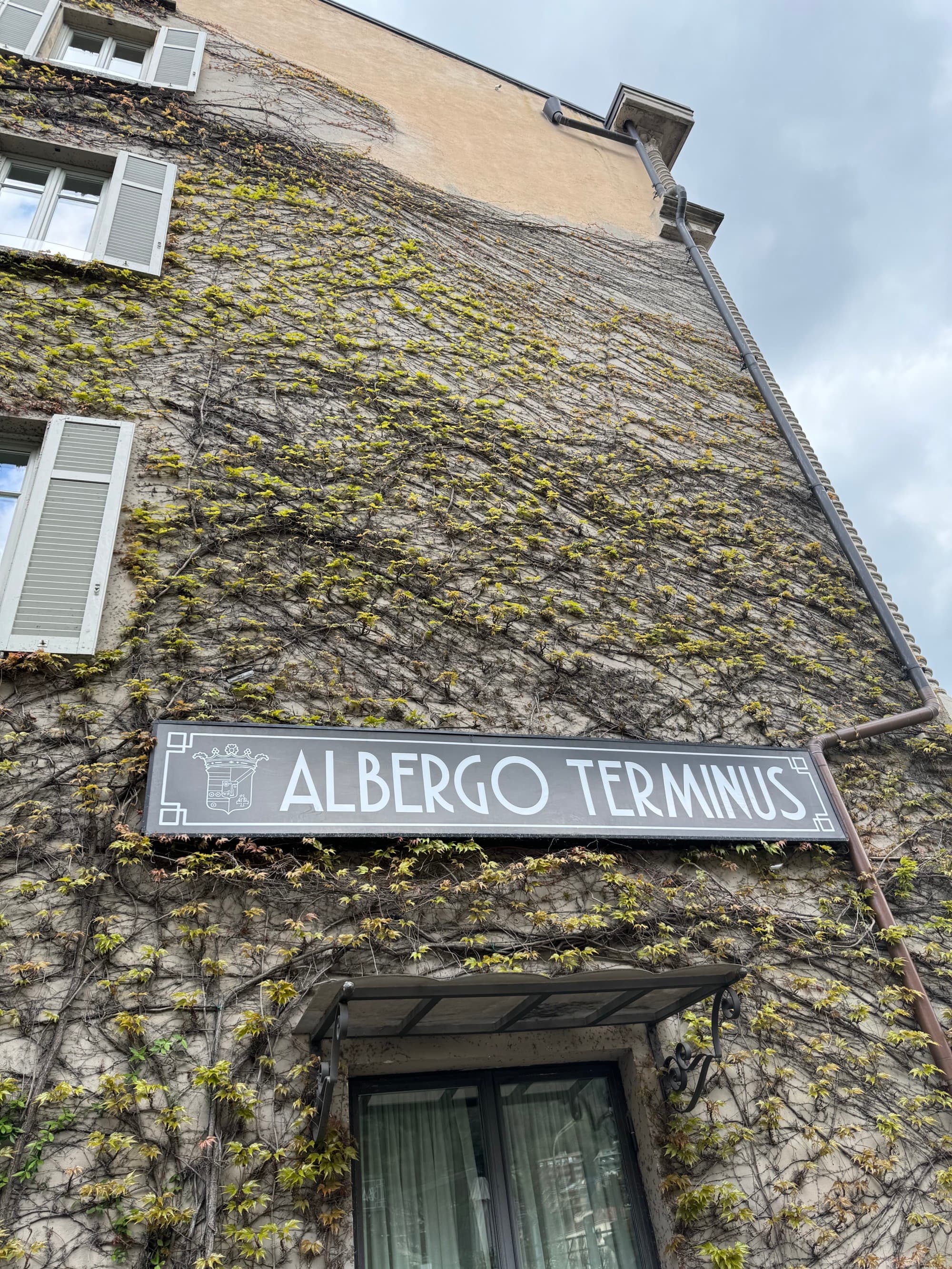 A photo of the corner of a stone house covered in leafless vines and showing an old street sign that reads "Albergo Terminus".