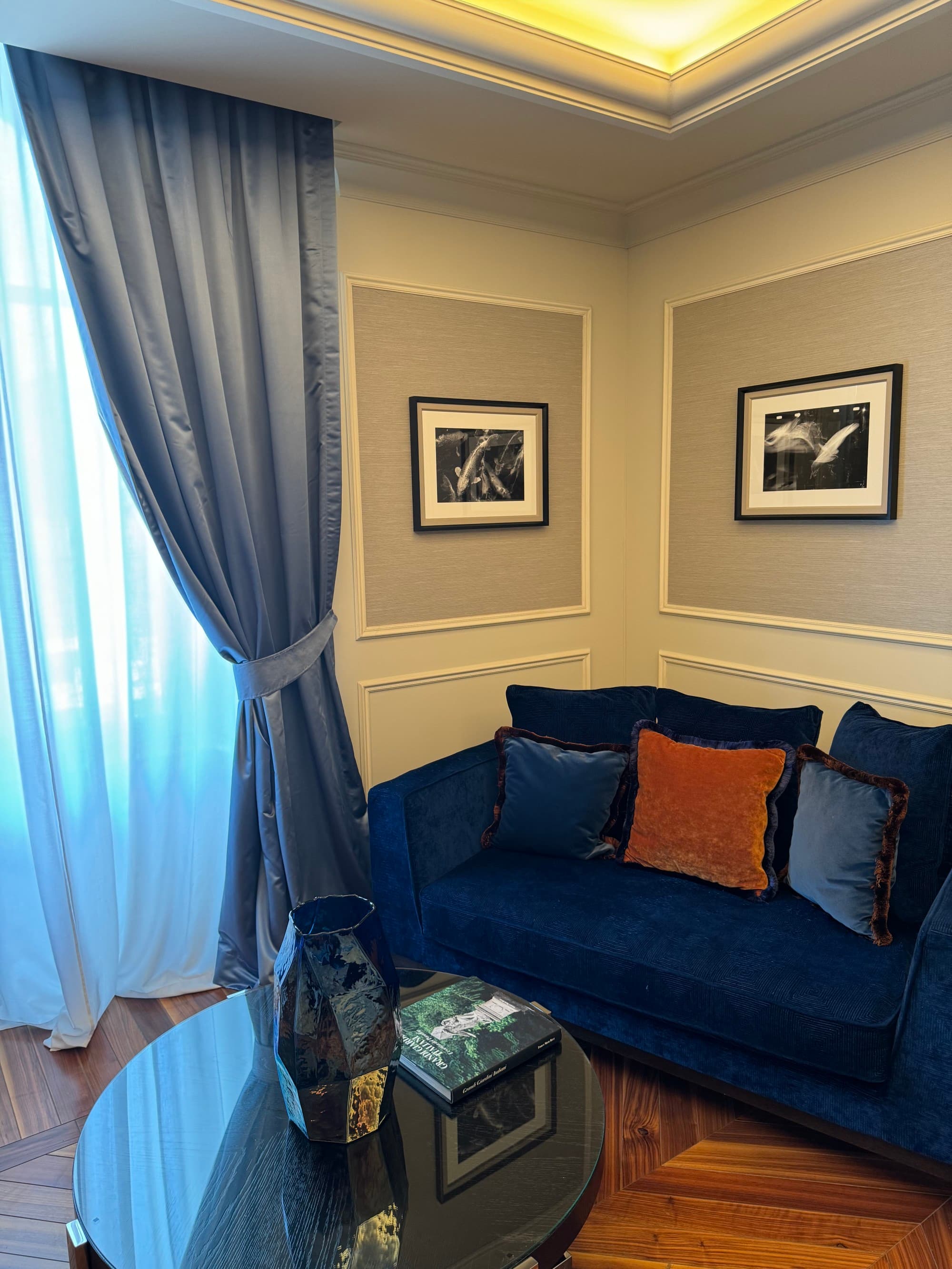 A sitting area with a deep blue couch, gray walls and a window letting light into the room with long blue curtains hanging on it.