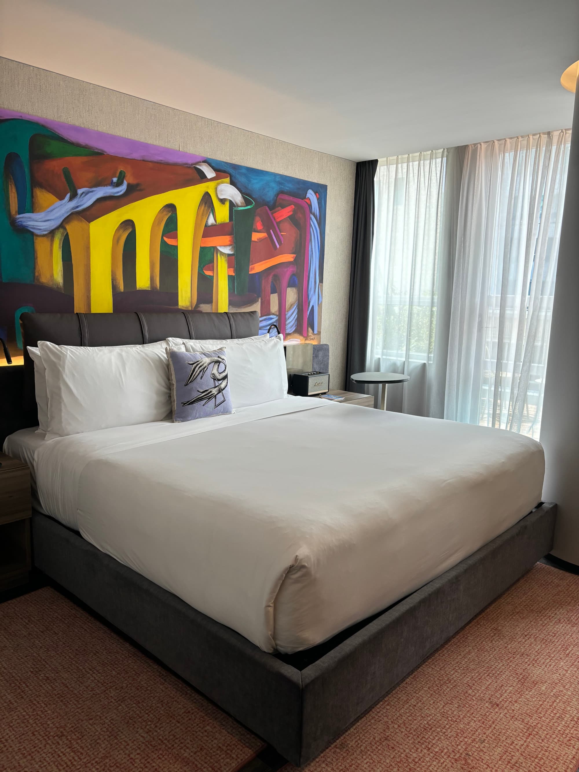 A queen-sized bed in one of the hotel's rooms with white sheets, and a colorful, abstract painting behind it.