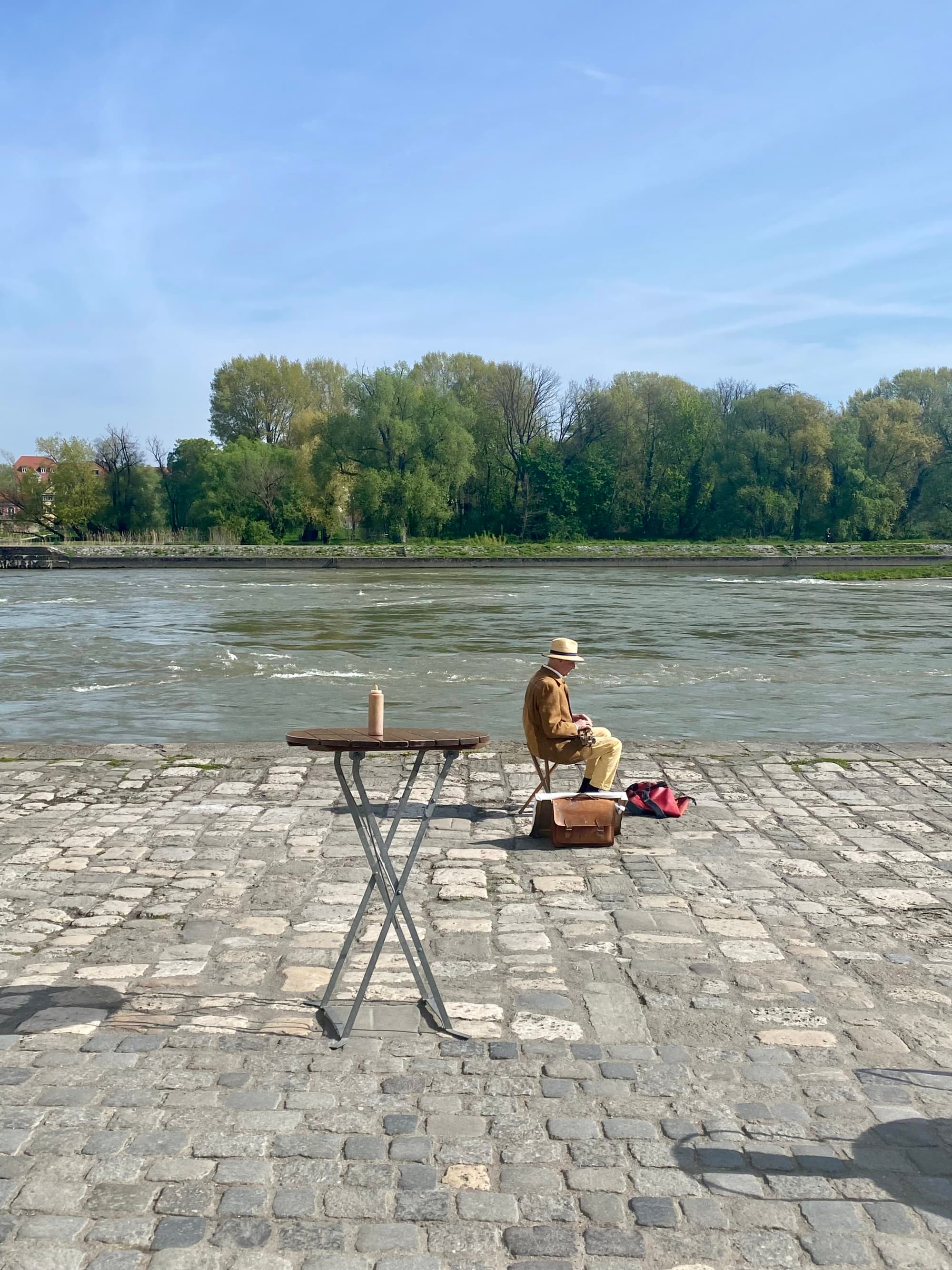 A local man sitting in a chair on a cobblestone shore to the Danube river, with trees in the background on a sunny day.