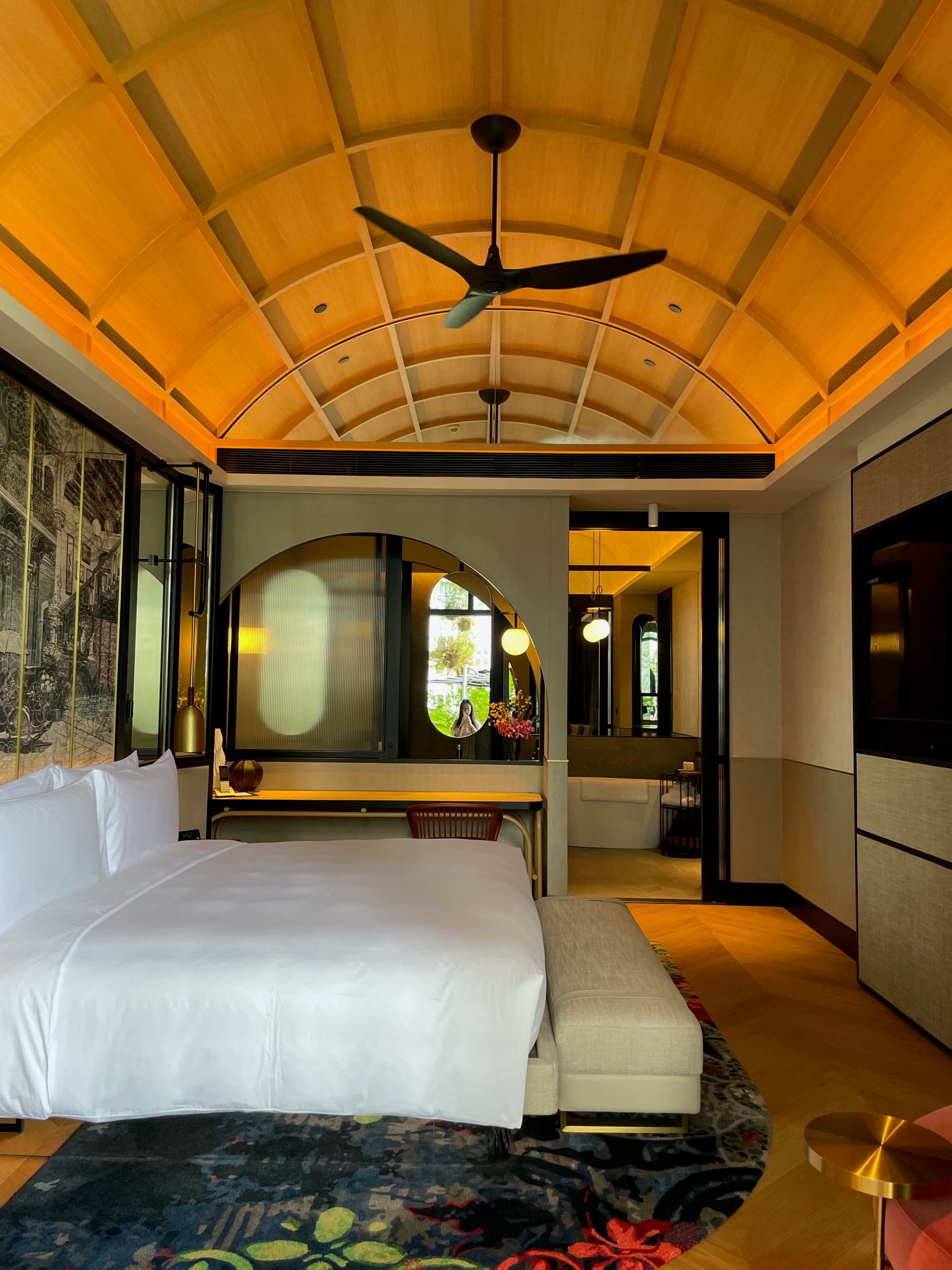 An elegant hotel bedroom with an arched ceiling, ceiling fan, large bed with white bedding and beige colored walls