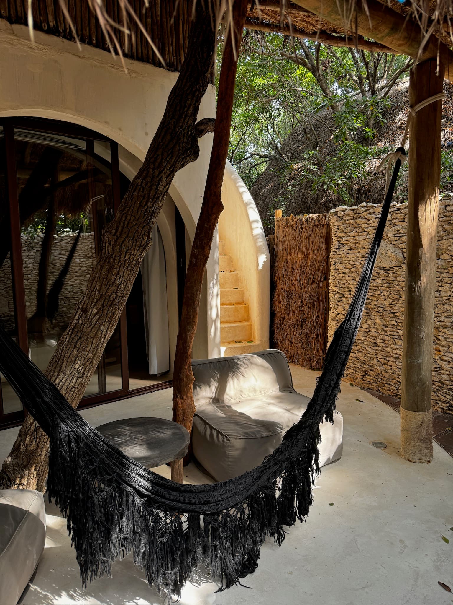 A black swing tied to trees, in front of a lounge chair and stone building with a staircase on the right in the background.