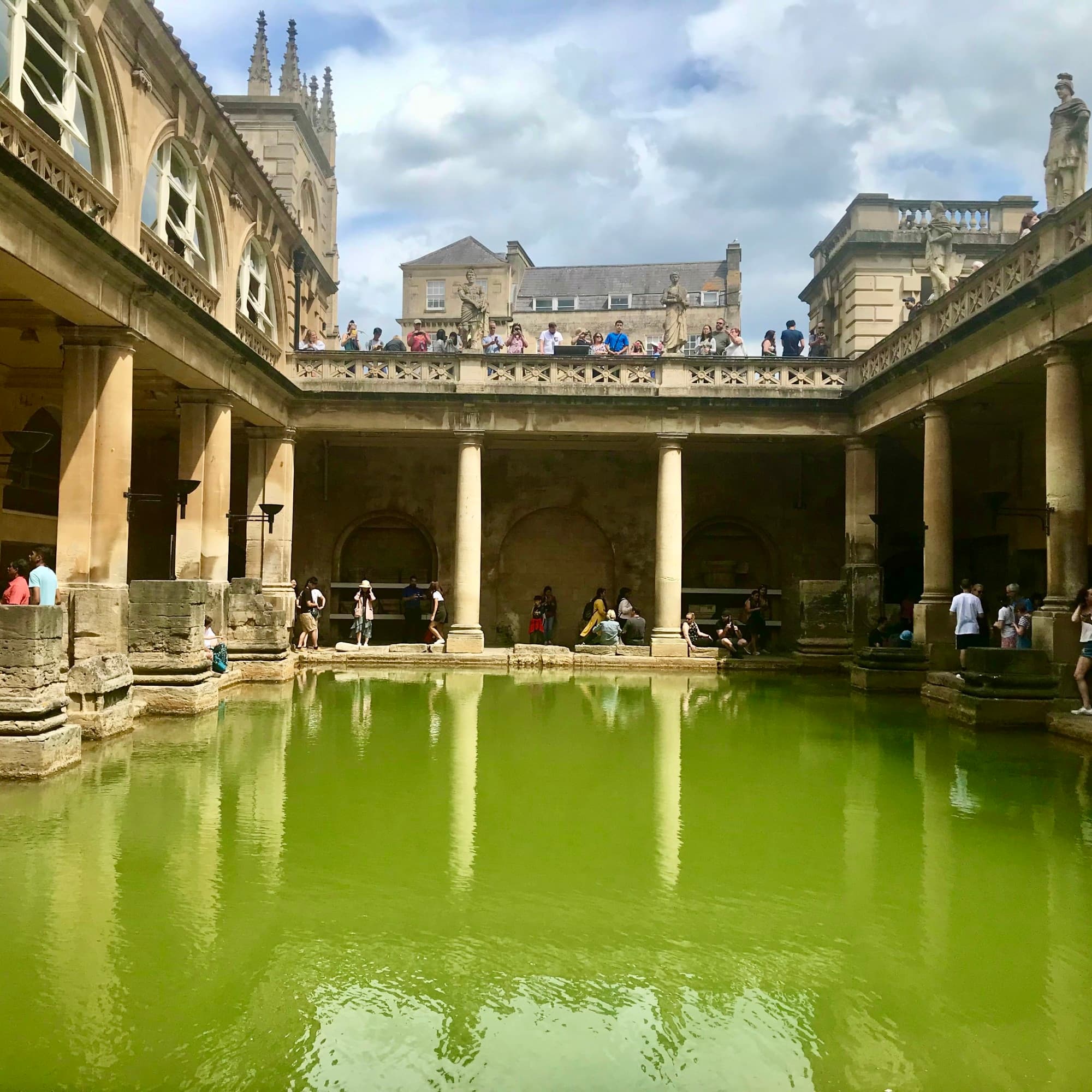 A green Roman bath surrounded by tourists and old stone architecture.