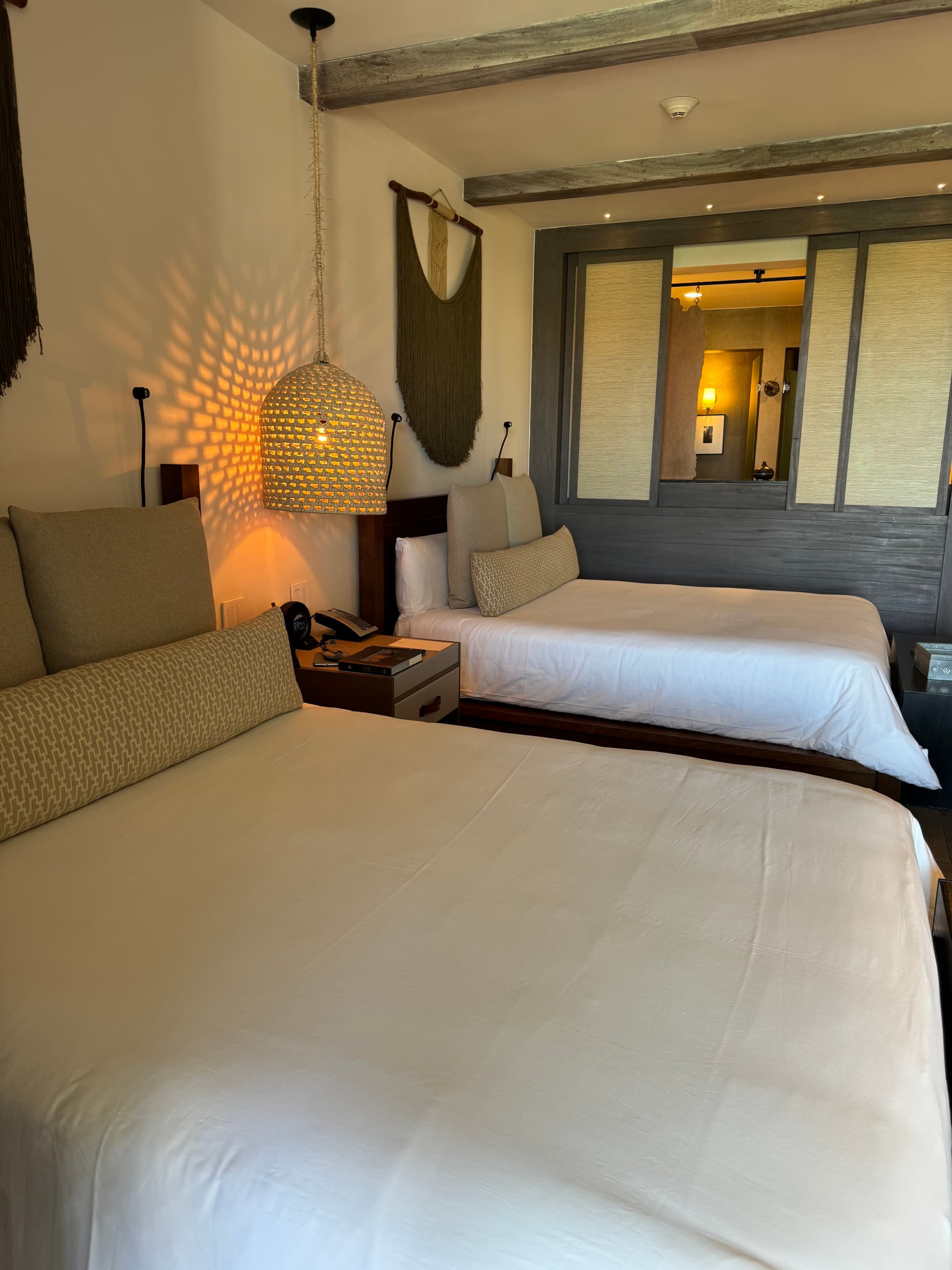 A hotel bedroom complete with two white beds, a wooden nightstand, a wicker light fixture hanging from the ceiling, sliding doors and art on the wall.