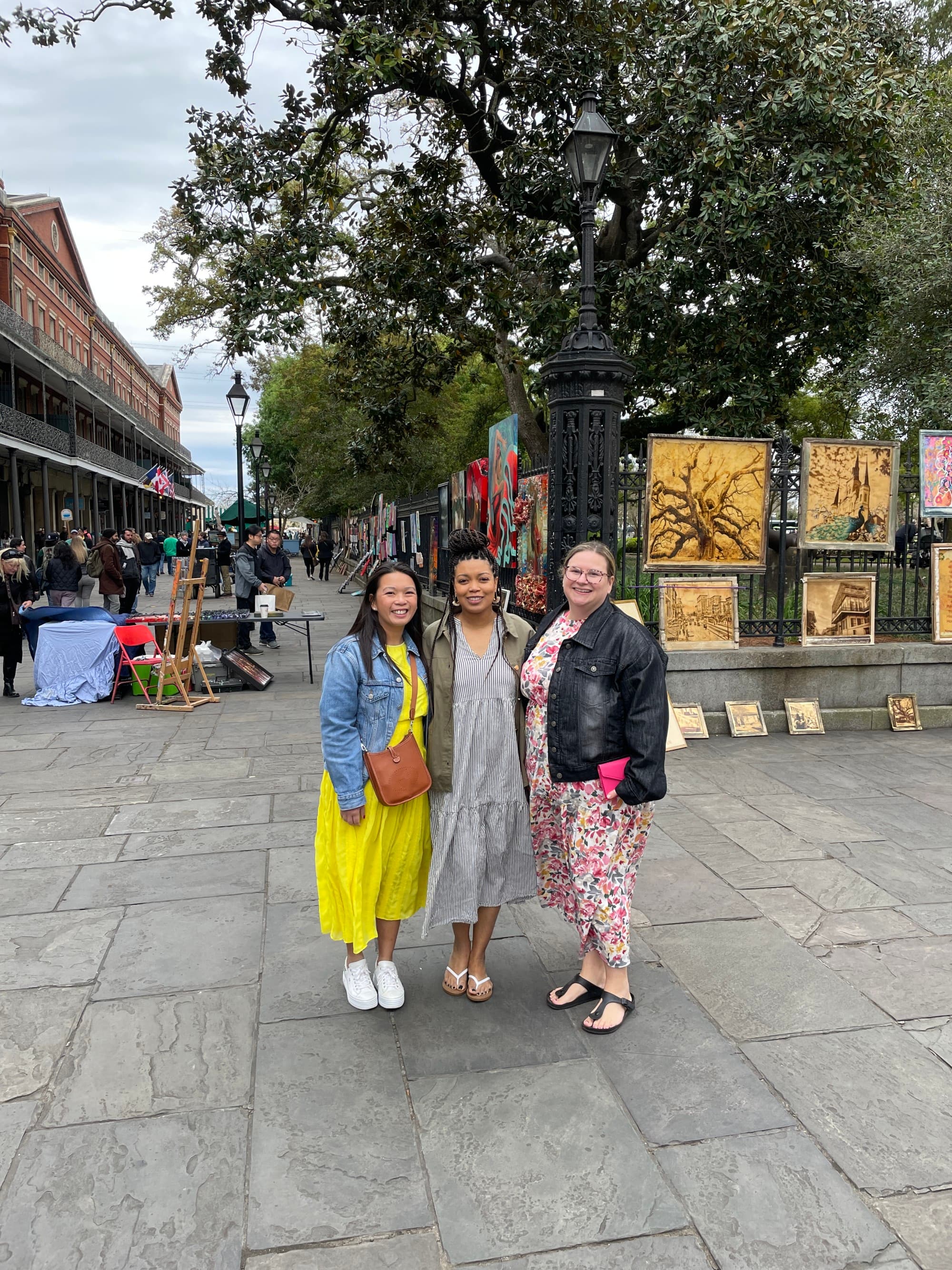 Three women posing in colorful dresses, on top of a stone road with metal fences showcasing artwork in the background.