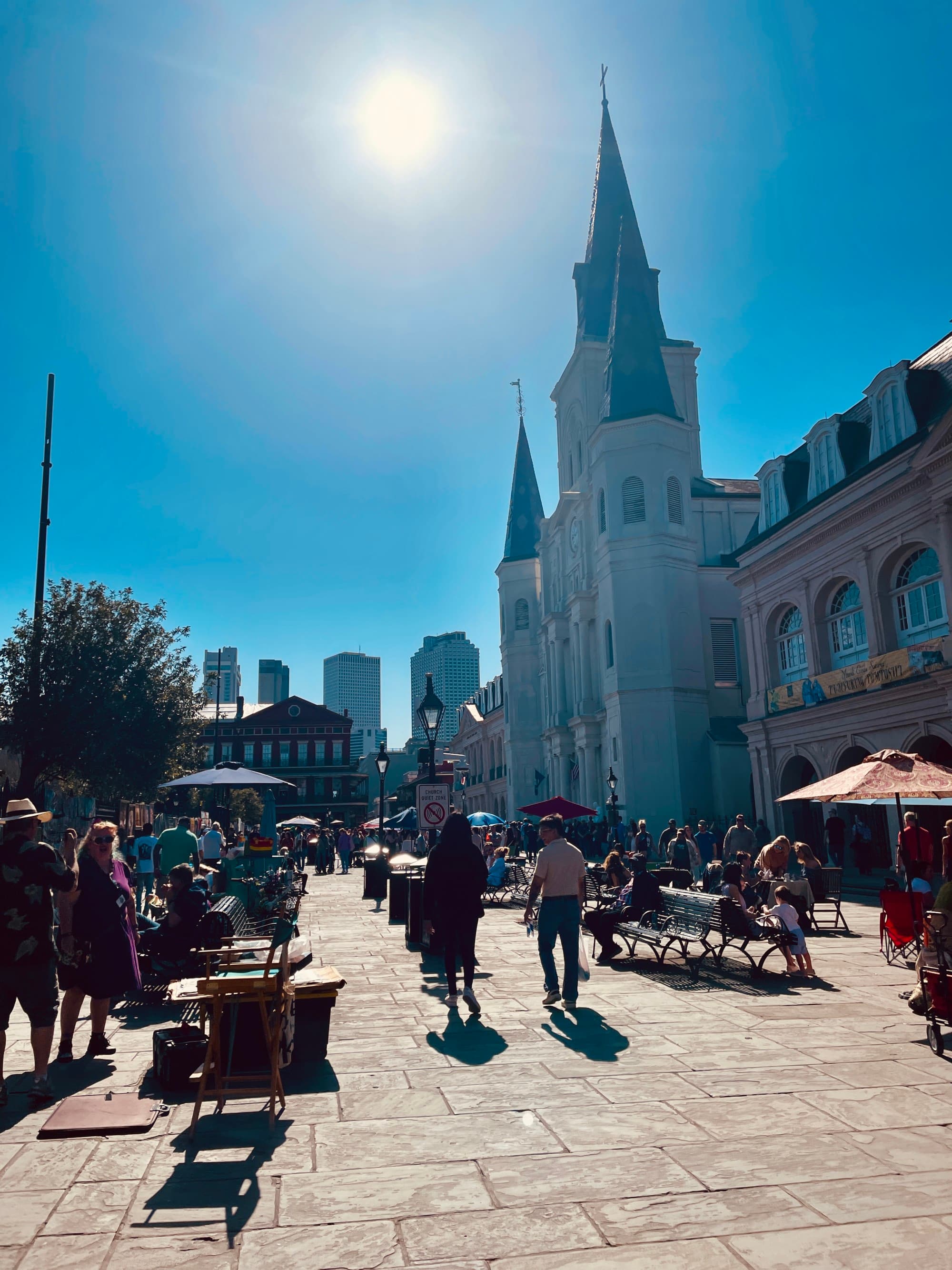 A view of people walking down a stone street with grand and old architecturally designed buildings under the beaming round sun.