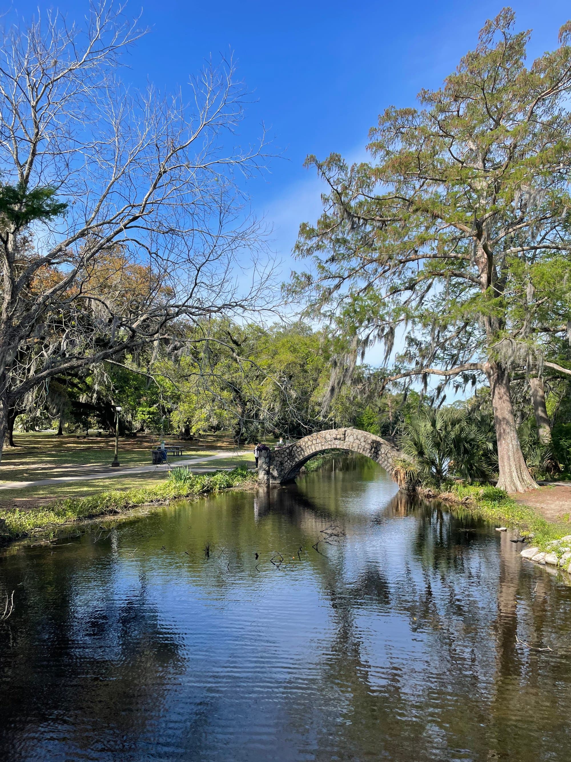 A calm lake with a stone arched bridge, trees, grass and a blue sky in the background.