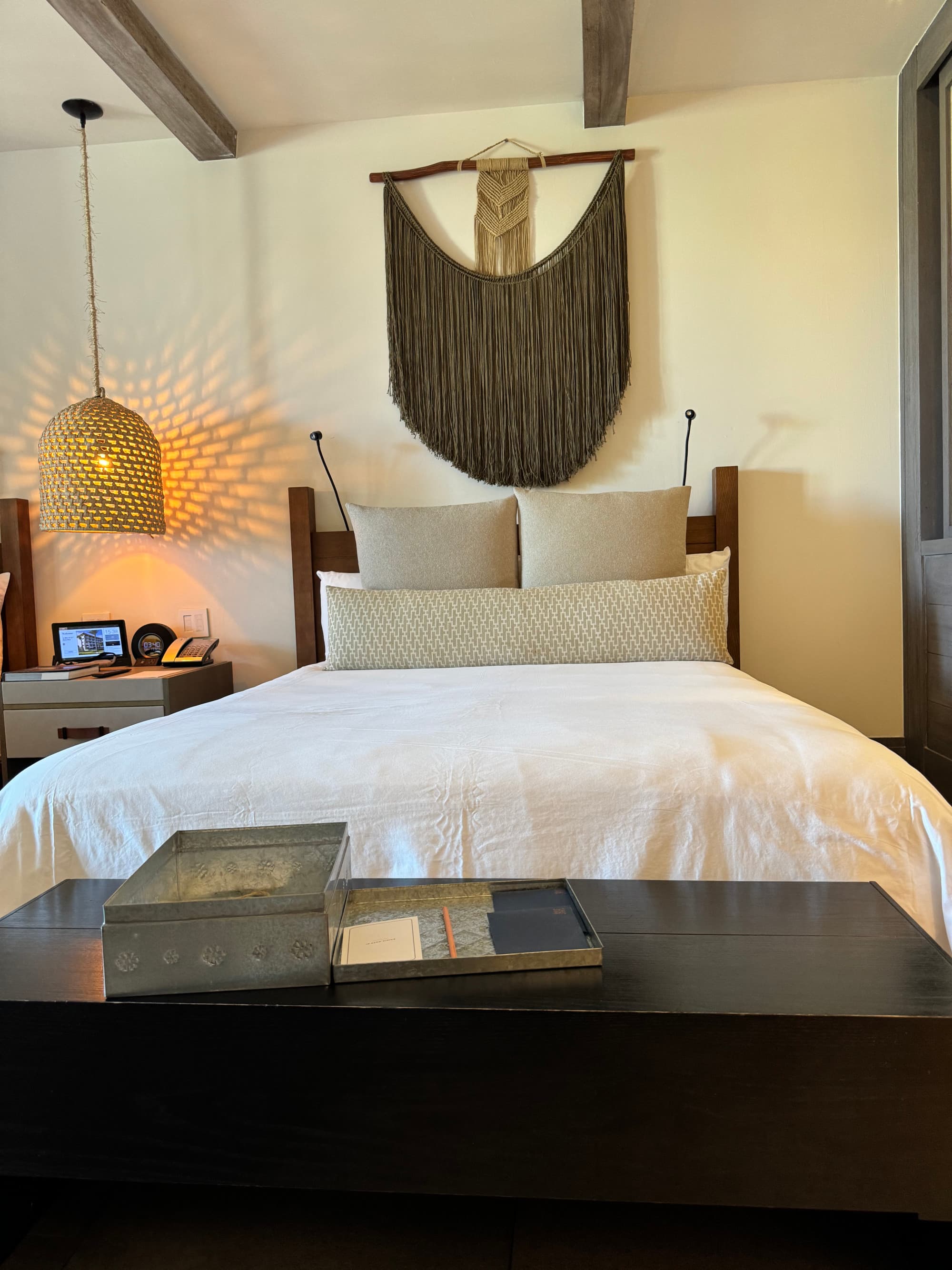 A white bed with a wooden table at the end of it with books on top of it. There is a wicker light hanging from the ceiling to the left of the bed and a decorative piece of artwork above the bed.