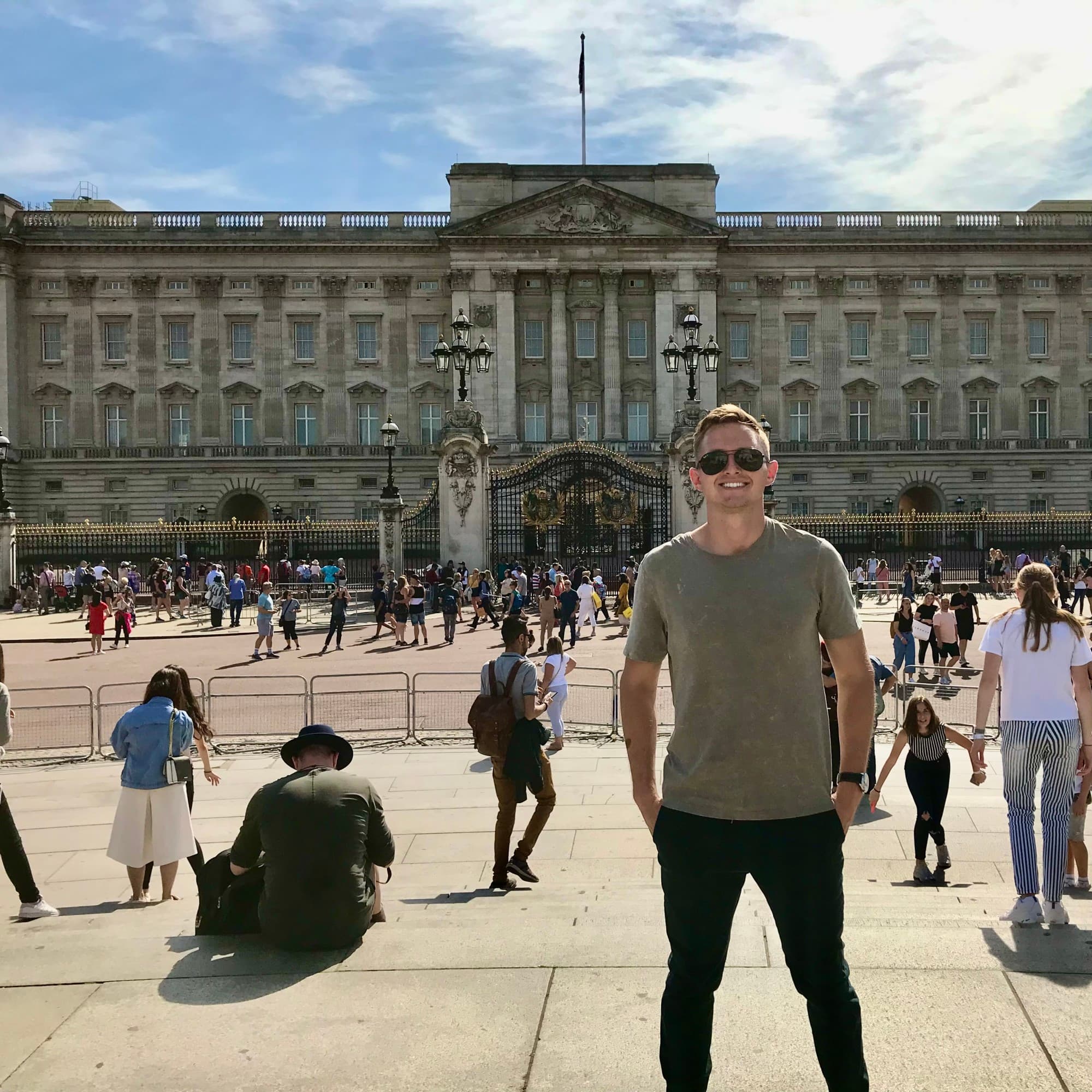 A man wearing sunglasses and a green t-shirt standing in front of Buckingham Palace surrounded by tourists.