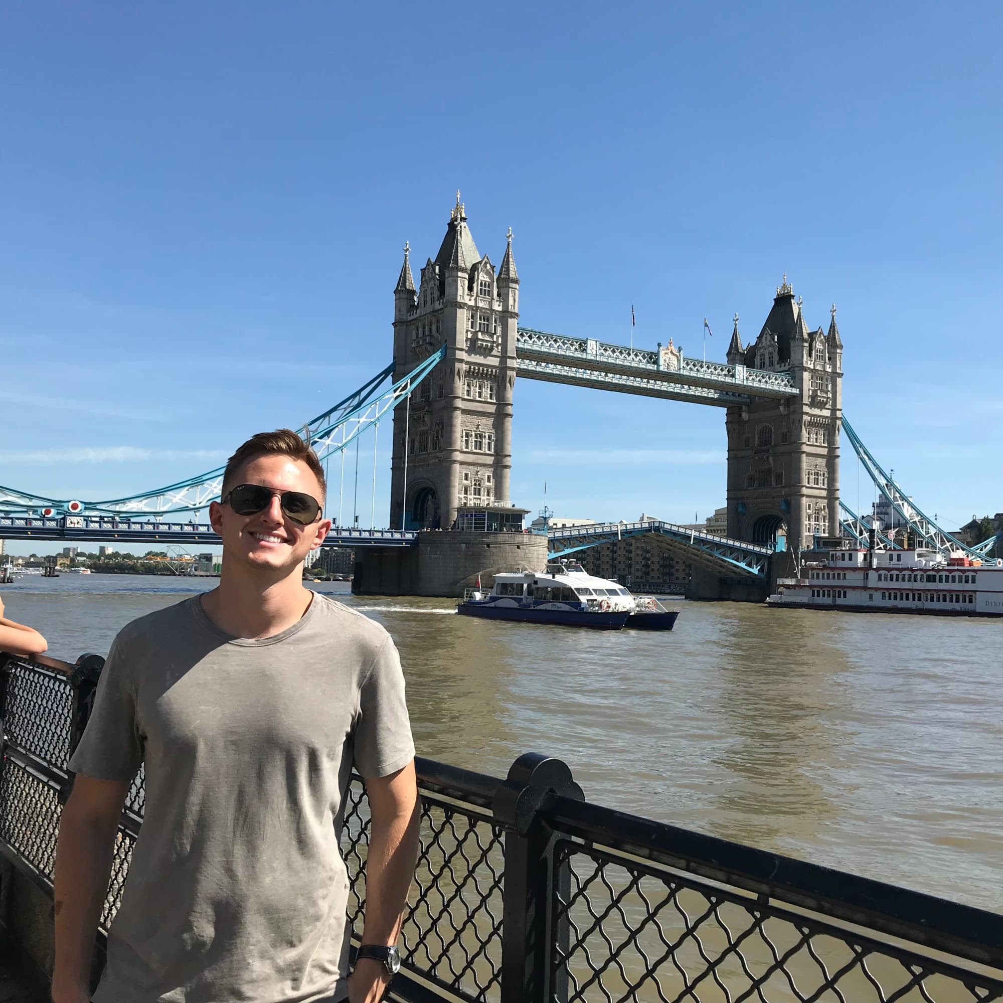 Joshua posing in front of the London bridge while wearing a grey t-shirt and sunglasses.