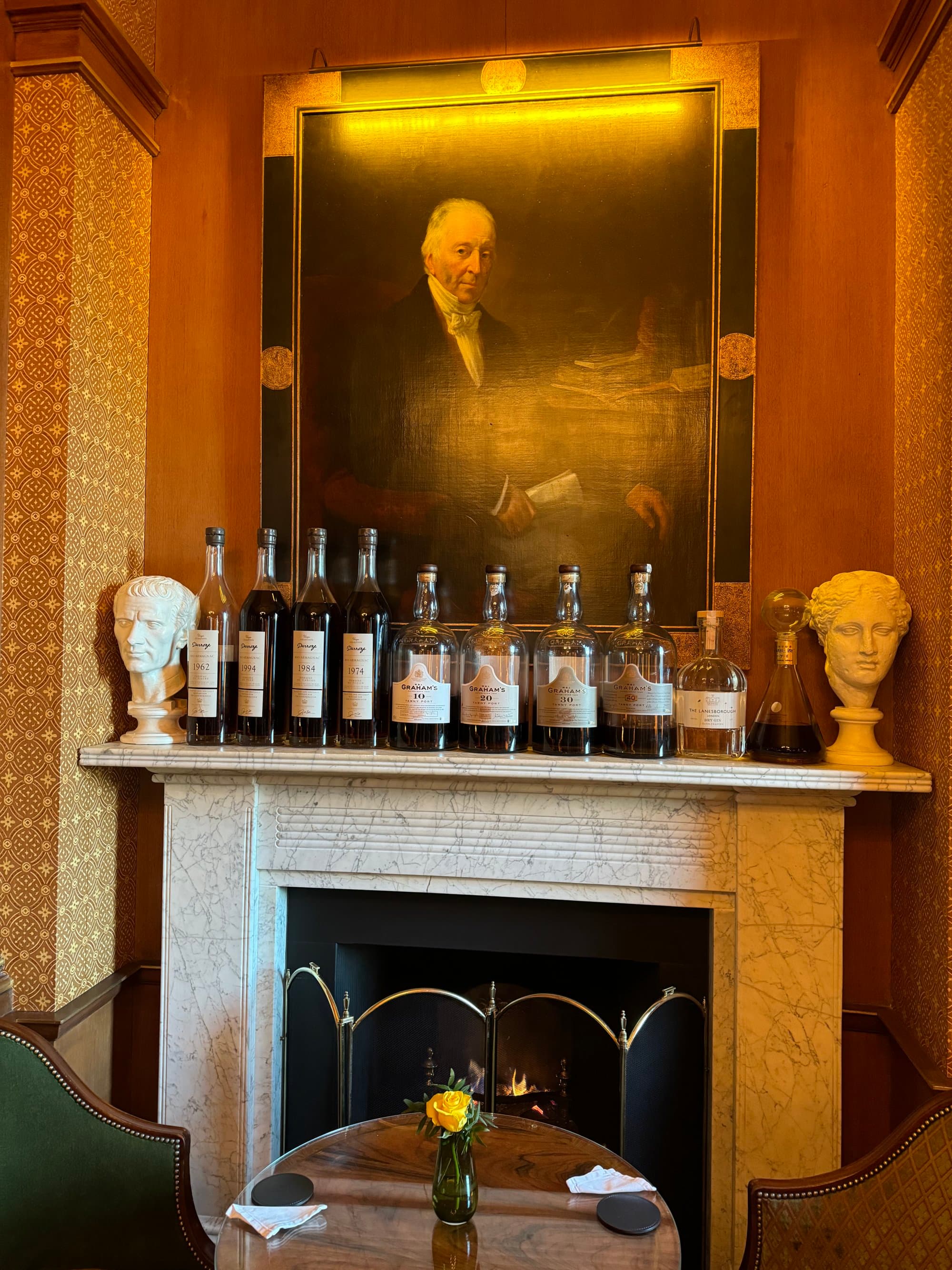 A picture of a fireplace with bottles of alcohol, busts, a painting of a man above or on it, and a round wooden table with two chairs in front of it.