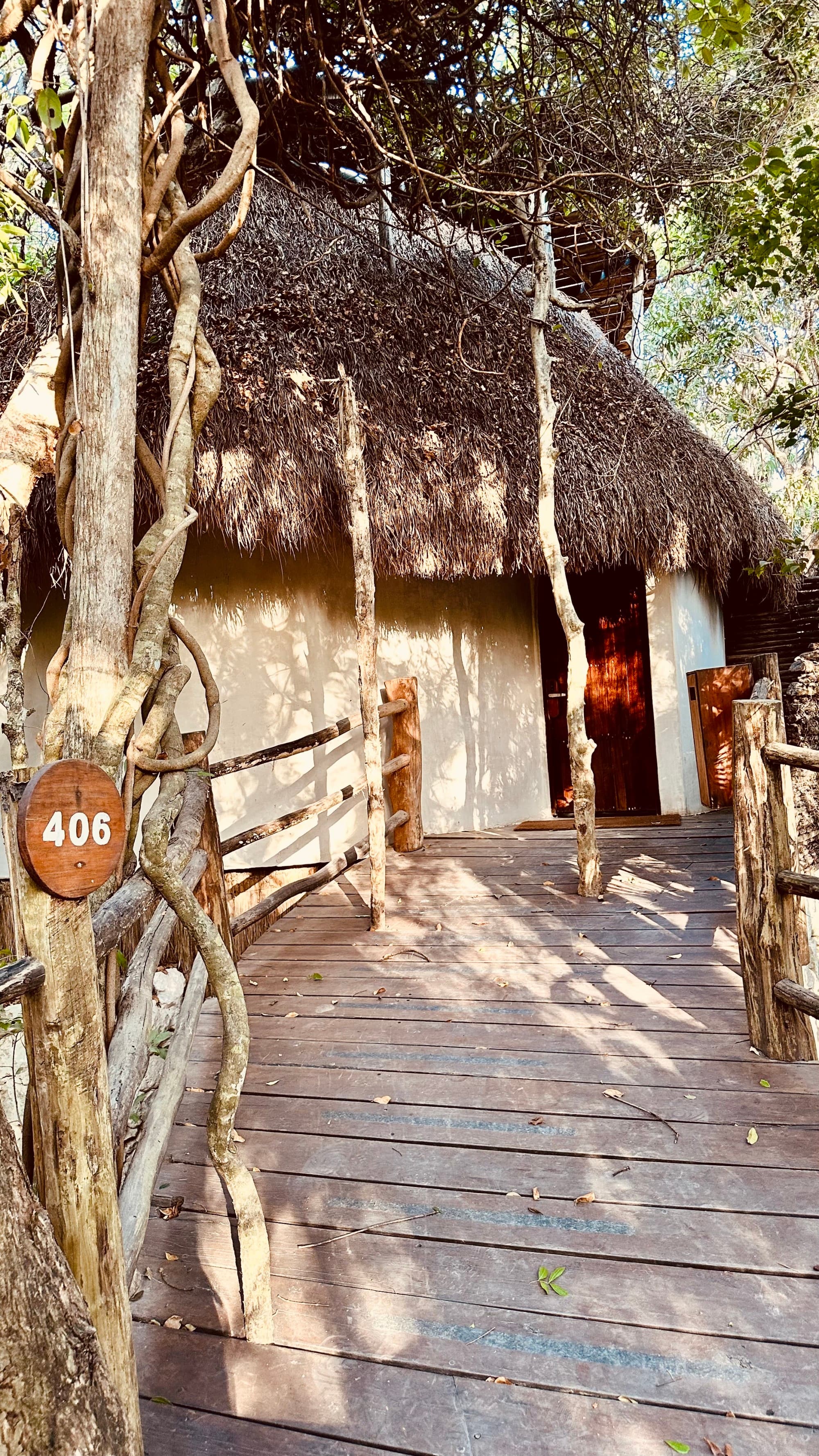 A wooden path leading to a hut with a straw roof, surrounded by trees and branches.