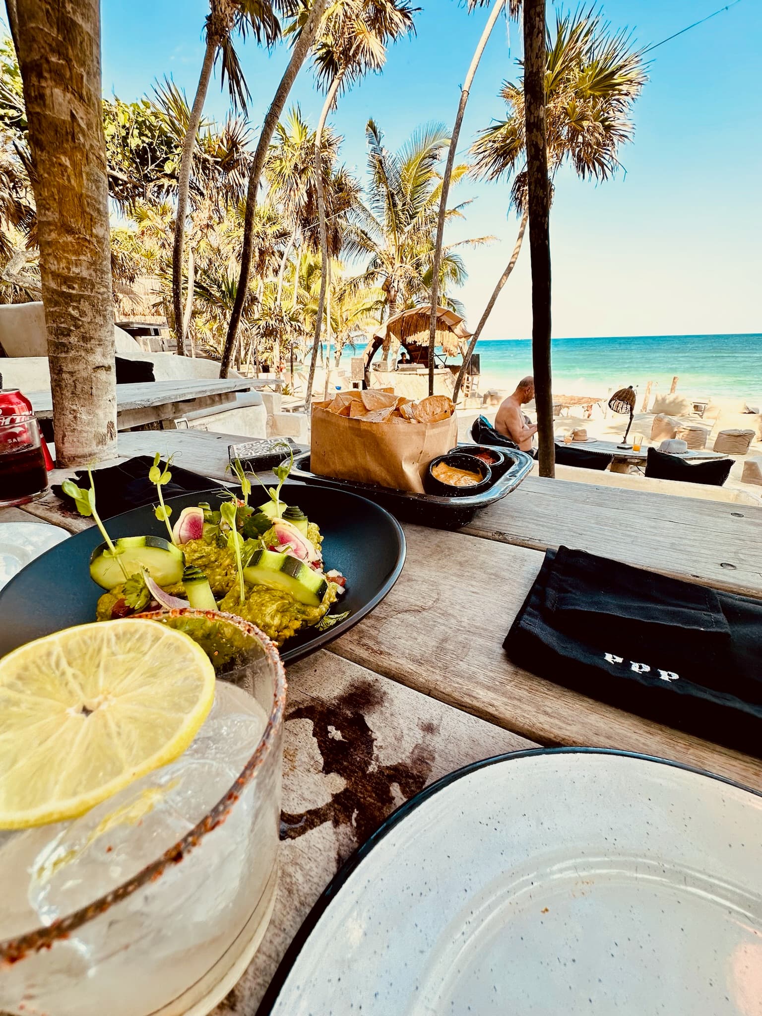 A wooden table with plates, colorful food, a beverage with a citrus slice and silverware on top of it. It's overlooking the beach, people lounging and the ocean, while surrounded by palm trees.